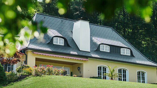 Felújított tetős családi ház, PREFA tetőfedő zsindely antracit színben, kerek tetőablakok (ökörszem ablak) és fehér kémény.