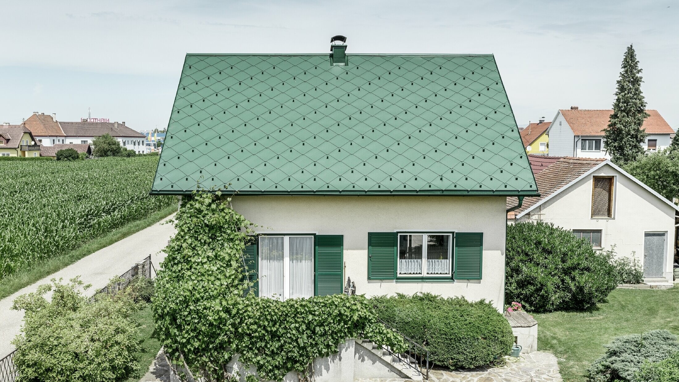 Klasszikus családi ház nyeregtetővel, mohazöld alumínium tetőfedéssel, zöld redőnyökkel. A tetőt PREFA 44 × 44-es tetőfedő rombuszok borítják P.10 mohazöld színben.