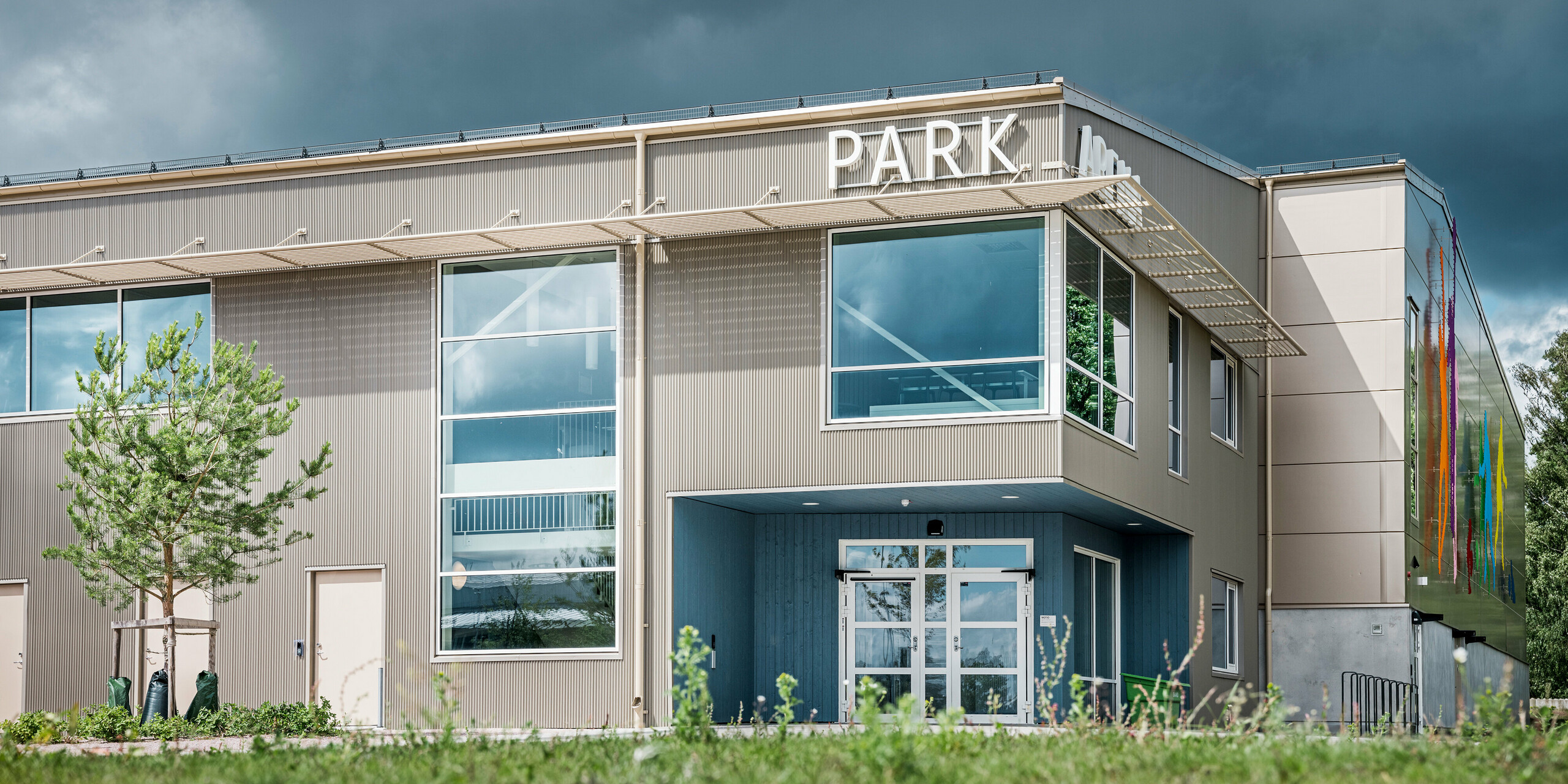 Eingangsbereich der Park Arena in Värnamo, Schweden, aus der Froschperspektive. Die Fassade ist mit dem markanten PREFA Zackenprofil in Bronze verkleidet, das für elegante Licht- und Schatteneffekte sorgt. Die modernen Glasfronten und der künstlerisch gestalteten Figuren an der Gebäudehülle betonen das innovative Design der Sporthalle.