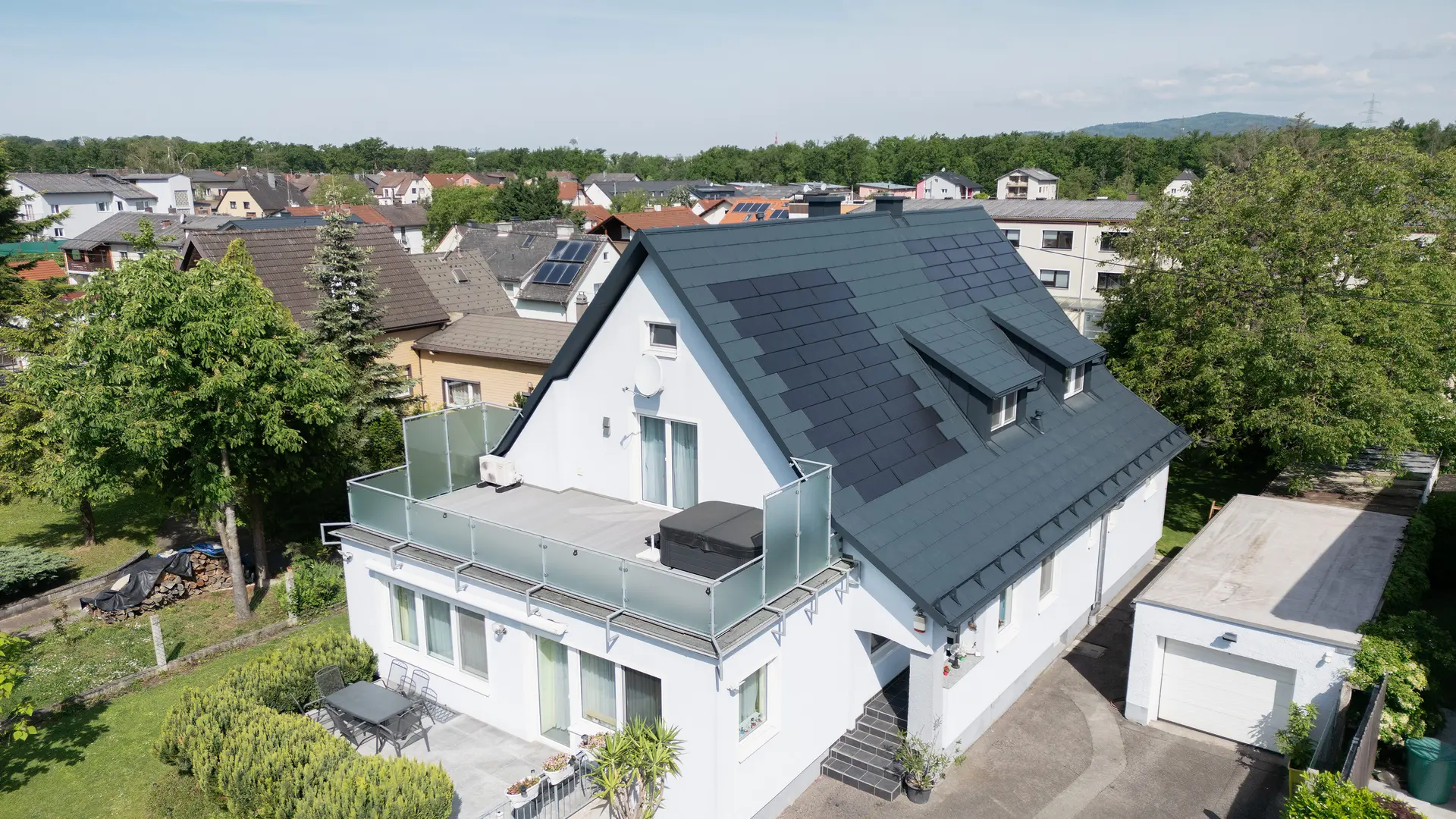 Madártávlatból egy családi ház a tetőfelújítás után. A tetőt most nagy PREFA napelemes tetőfedő panelek és a PREFA R.16 Classic elem borítják, P.10 antracit színben. A napelemes tetőfedő panelek egyenletesen vannak elosztva, és modern, fenntartható megoldást nyújtanak. Az épület most modern és környezetbarát benyomást kelt. Az előtérben látható erkély üvegkorláttal rendelkezik, és az épület felső emeletén található.