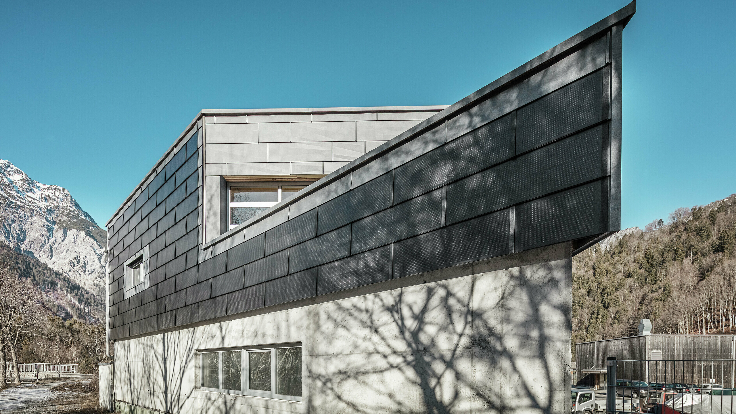 Das Firmengebäude der Spenglerei RÜMA in Vorarlberg beeindruckt mit seiner modernen Fassade aus PREFA Solardachplatten und Sichtbeton. Umgeben von Bergen und Bäumen, deren Schatten sich auf der Wand abzeichnen, steht es unter einem klaren, blauen Himmel. Die spitze Gebäudeecke und die PREFA Fassadenpaneele FX.12 betonen das markante Erscheinungsbild zusätzlich.