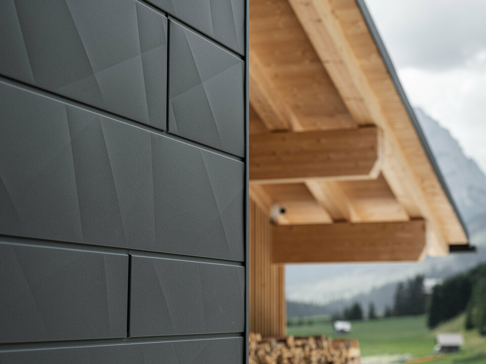 Közelkép a PREFA Siding.X homlokzatpanelekről P.10 fekete színben a Casa Hoffe épületén Sappada városában, Olaszországban. Az alumínium homlokzati rendszer modern, absztrakt kialakításban került alkalmazásra, amely vonzó kontrasztot képez a tetőszerkezet látható faelemeivel. Az innovatív homlokzati megoldás zökkenőmentesen illeszkedik az alpesi környezetbe, és a funkcionalitás, a tartósság és az esztétikus építészeti kultúra ötvözetét képviseli.