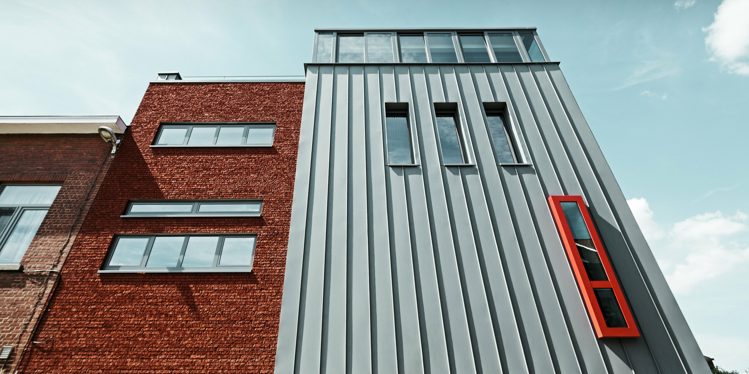 Stilvolle Kombination aus traditioneller Ziegelfassade und innovativer PREFALZ Aluminiumfassade in Patinagrau. Die markante vertikale Stehfalzstruktur trifft auf ein kräftig rotes Akzentfenster und klare Fensterlinien. Das langlebige Aluminium schützt dauerhaft vor Witterungseinflüssen und verleiht dem Gebäude einen zeitlos modernen Charakter.