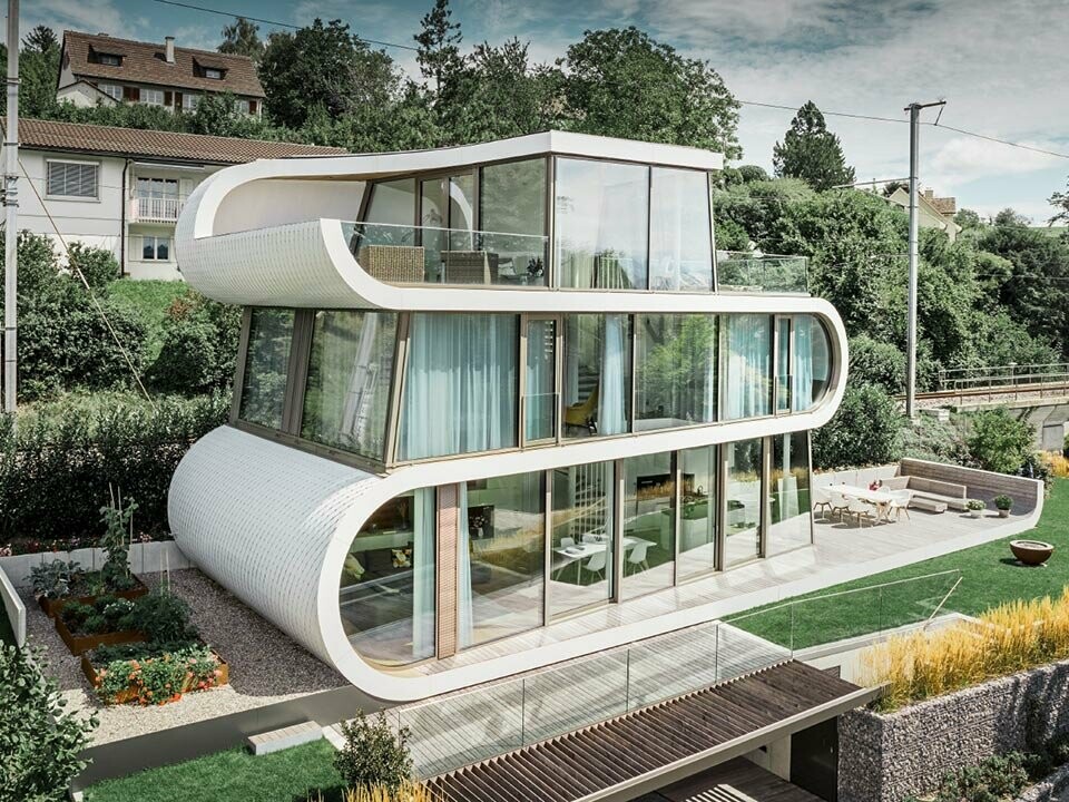 Fénykép a Zürichi-tónál épült modern Flexhouse-ról, amely szokatlan épületformájával vonzza magára a tekintetet. Az ívelt szintelválasztók fehér szalagként húzódnak végig az épületen. A kerek homlokzati elemeket tisztafehér színű PREFA alumíniumzsindellyel burkolták.