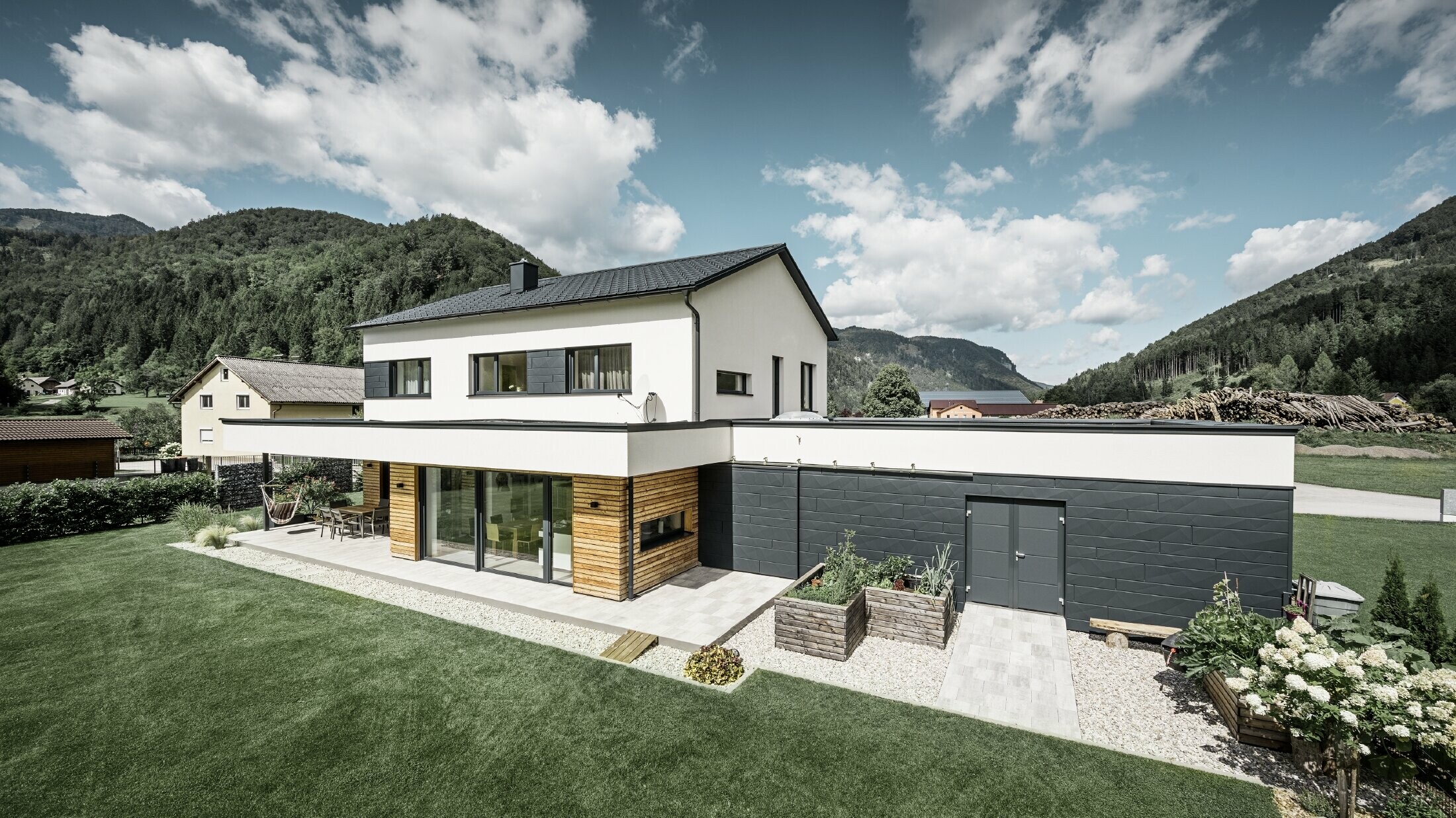Modern családi ház fehér vakolattal és PREFA Siding.X P.10 antracit színben, idilli vidékkel körülvéve. A sötét homlokzat kontrasztot alkot a világos környezettel. A classic elemek és a tető vízelvezetése teszi teljessé a tartós kialakítást.