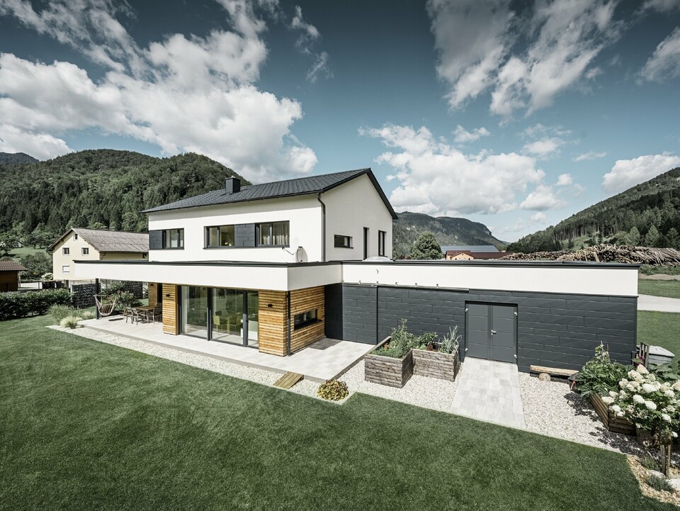 Modern családi ház fehér vakolattal és PREFA Siding.X P.10 antracit színben, idilli vidékkel körülvéve. A sötét homlokzat kontrasztot alkot a világos környezettel. A classic elemek és a tető vízelvezetése teszi teljessé a tartós kialakítást.