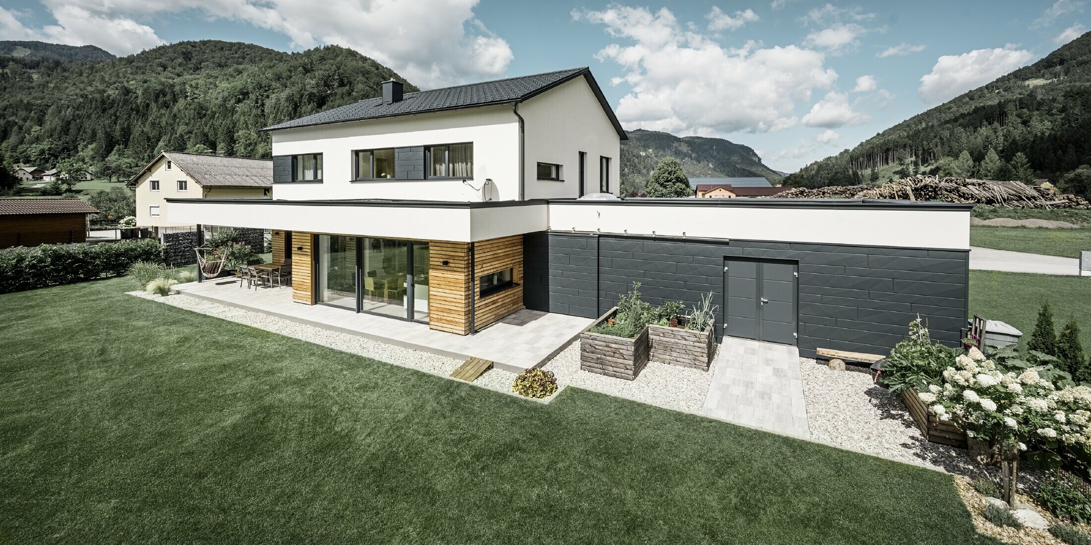 Modern családi ház fehér vakolattal és PREFA Siding.X P.10 antracit színben, idilli vidékkel körülvéve. A sötét homlokzat kontrasztot alkot a világos környezettel. A classic elemek és a tető vízelvezetése teszi teljessé a tartós kialakítást.
