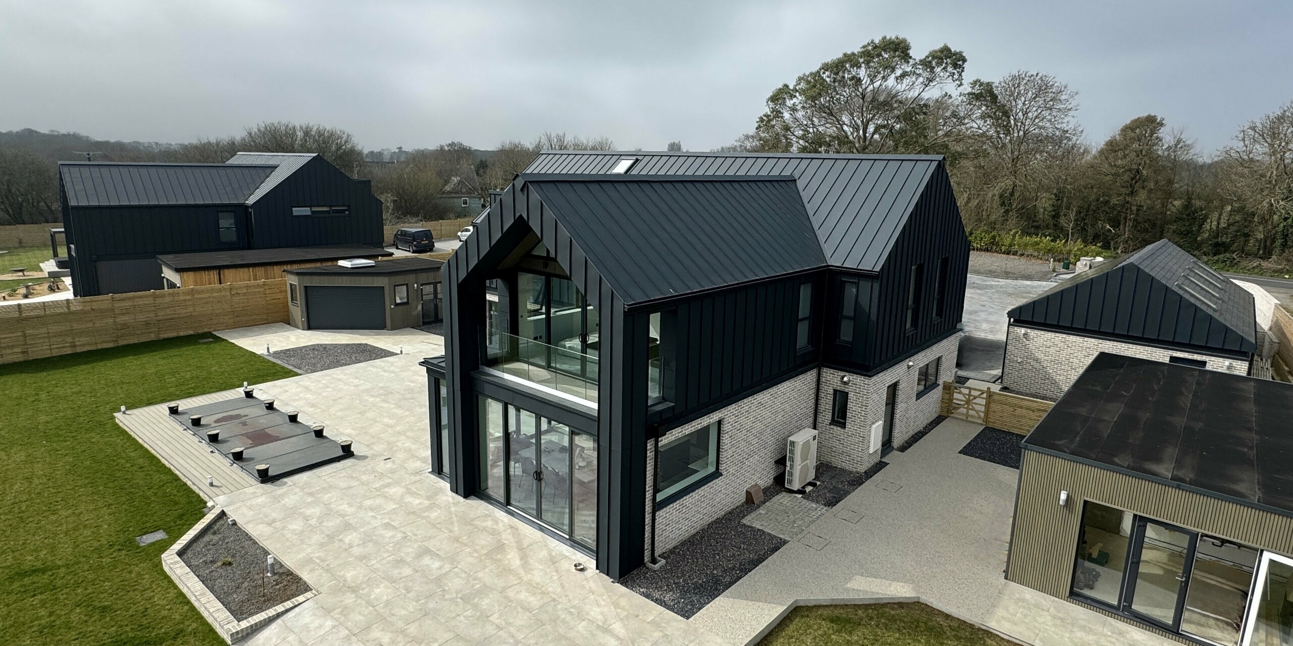 Ansicht eines modernen Einfamilienhauses mit PREFALZ Dach und Fassade in P.10 Dunkelgrau, kombiniert mit heller Steinverkleidung im Erdgeschoss. Auffällig sind die großen Glasfronten, die für viel Licht sorgen. Im Vordergrund eine großzügige Terrasse mit klarer Linienführung, im Hintergrund weitere Gebäude des Ensembles Bullenmead. Die Aluminiumoberflächen sind langlebig, korrosionsbeständig und ideal für die Küstenlage. Das Zusammenspiel von dunklem Dach, Glas und Naturstein unterstreicht die elegante Architektur.