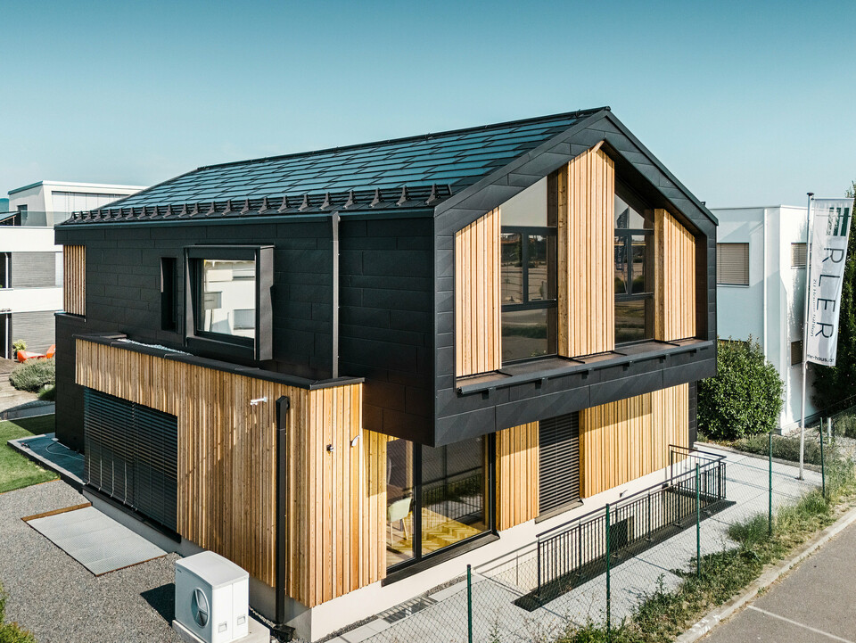 Az Autark mintaház elülső része a Blaue Lagune előre gyártott házak központjában, Vösendorfban (AT). A tető fekete PREFA napelemes Classic elemekkel és R.16-os Classic elemekkel van fedve, kiváló minőségű P.10 bevonattal. A homlokzat függőleges faelemek és PREFA Siding.X P.10 fekete színű elemek kombinációjából áll, ami modern és fenntartható jelleget kölcsönöz az épületnek. A kép jobb szélén az Erler Bau GmbH zászlaja lobog.