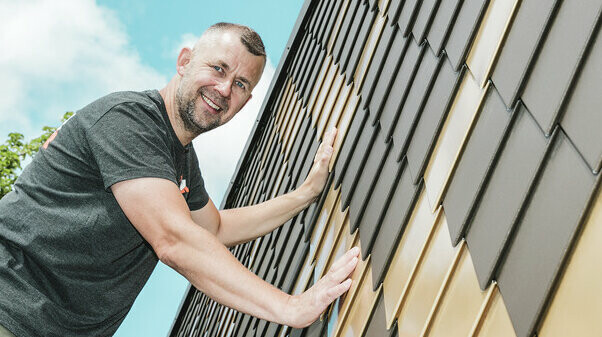 PREFA Handwerker mit grauem T-Shirt an einer PREFA Rauten-Fassade in Braun und Mayagold.