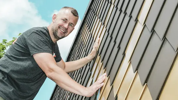 PREFA Handwerker mit grauem T-Shirt an einer PREFA Rauten-Fassade in Braun und Mayagold.