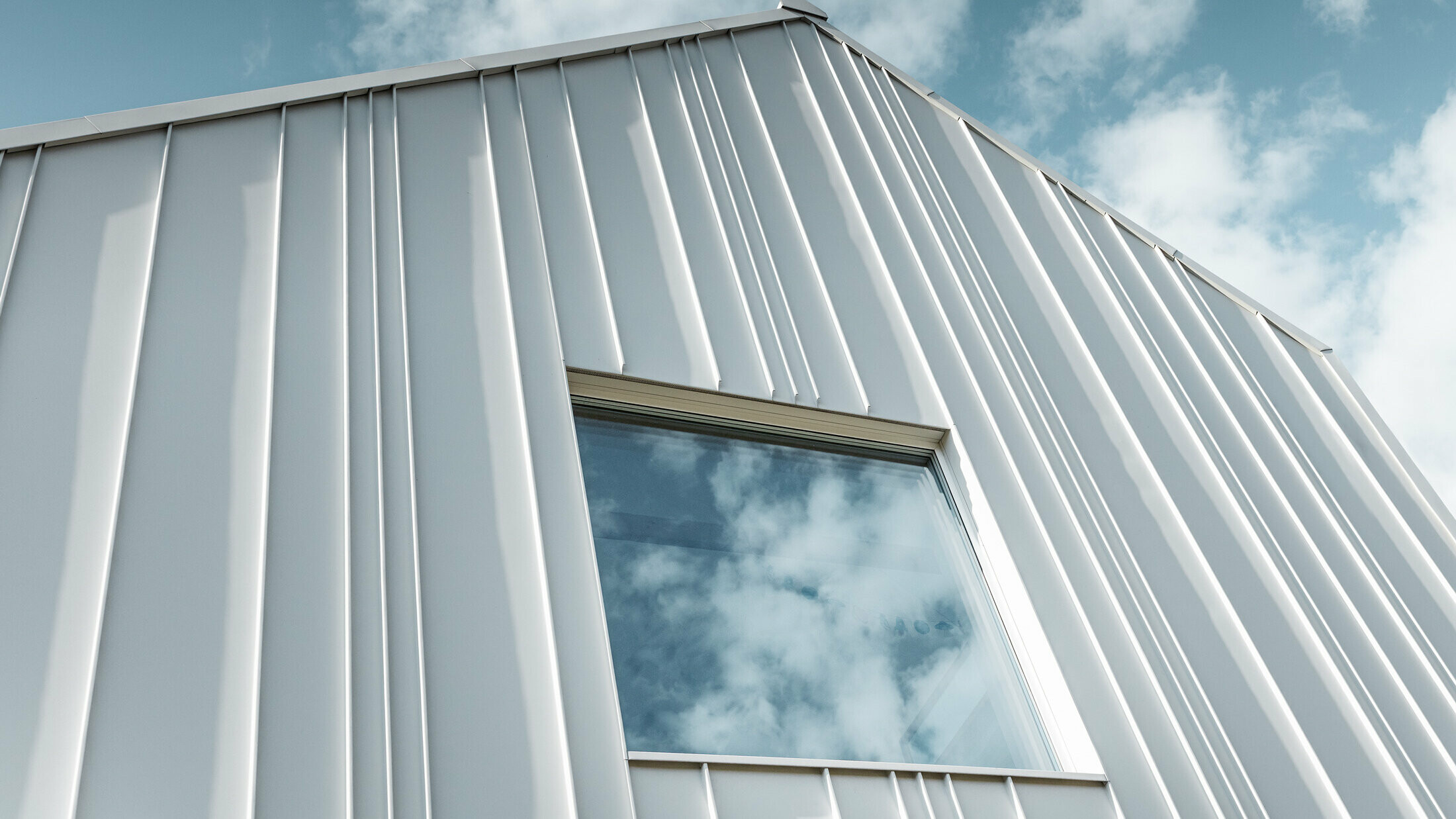 Fehér PREFALZ elem különböző sarniszélességekkel és egy ablakkal kialakított homlokzaton lévő derékszögű állókorcként