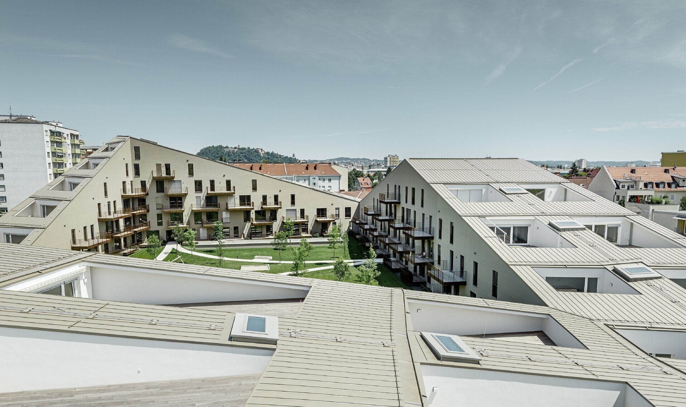 hosszasan elnyúló lakótelep Grazban, alacsony hajlású tetősíkokkal, beépített teraszokkal és nagy üvegfelületekkel, egyedi, bronz metál Prefalz korcolt lemezfedéssel