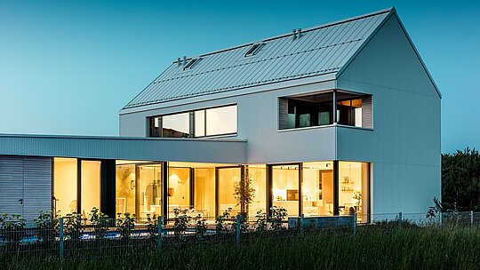 Das Bild zeigt das moderne Einfamilienhaus in Freistadt aus einer Perspektive, die das harmonische Zusammenspiel von Architektur und Natur betont. Die markante PREFALZ-Fassade in Reinweiß zieht sofort die Aufmerksamkeit auf sich, während die großzügigen Glasflächen den Innenraum offen und einladend wirken lassen. Das steile Satteldach, ebenfalls mit PREFALZ-Aluminium gedeckt, sorgt für einen dynamischen Abschluss und schützt das Gebäude vor Witterungseinflüssen. Die stilvolle, minimalistische Gestaltung wird durch die warmen Holzelemente am Balkon und an der Terrasse akzentuiert, was für eine angenehme Kontrasterfahrung sorgt. Das Gebäude ist nicht nur ästhetisch ansprechend, sondern auch funktional und nachhaltig dank der langlebigen und pflegeleichten PREFALZ-Aluminiumprodukte.