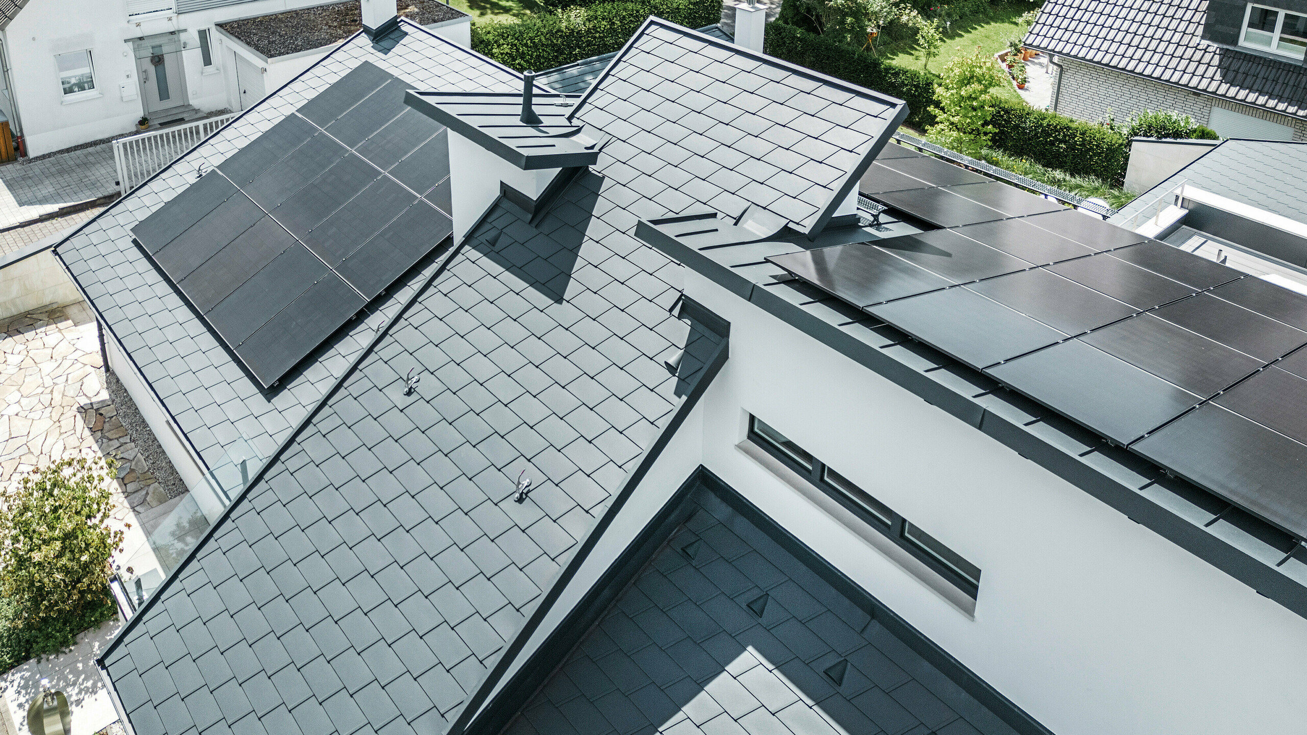 Modern családi ház Dortmundban, fejlett fotovoltaikus rendszerrel, amely DS.19 tetőzsindelyből és P.10 antracit színű PREFALZ állóhomlokzati tetőfedésből álló PREFA tetőszerkezetre van felszerelve. A PV-rendszer a tetőzsindelyeknél Vario szolárkonzolok, az állófóliáknál pedig PREFALZ Vario szolárkonzolok segítségével lett felszerelve. A napelemes rendszer hatékony energiatermelést biztosít megújuló energiaforrásokból. Az igényes tetőszerkezet optimális egyensúlyt kínál az esztétika és a funkcionalitás között.