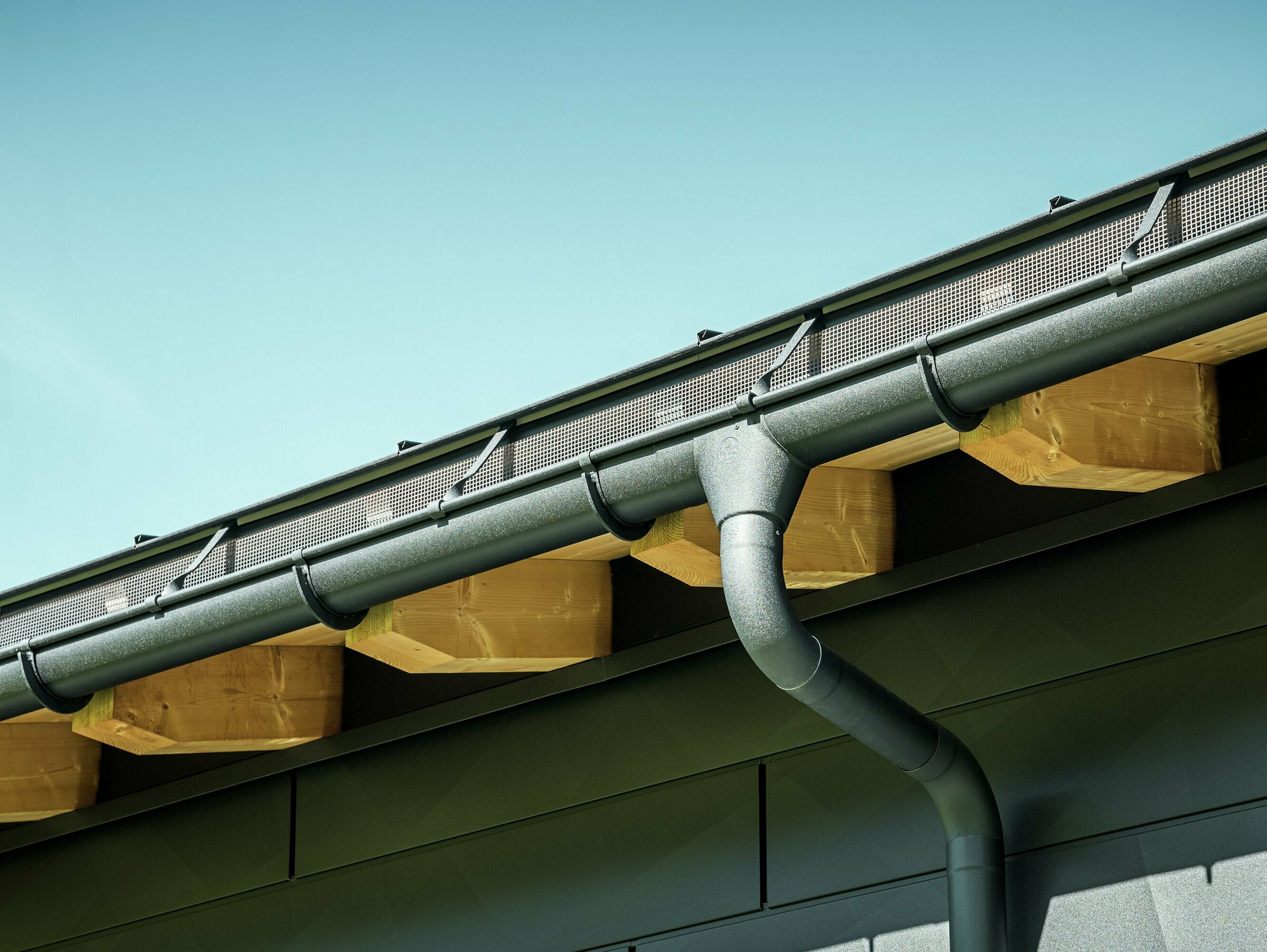 PREFA függesztett ereszcsatorna csatornatartóval és túlnyúló szegéllyel, P.10 fekete Közelkép egy modern családi ház homlokzatáról Ascharinában, Svájcban. Az épület oldalait a jellegzetes PREFA Siding.X homlokzatpanelek borítják P.10 fekete színben. Színben illeszkedik hozzá a PREFA tetővízelvezető rendszer függesztett ereszcsatornával és lefolyócsővel.