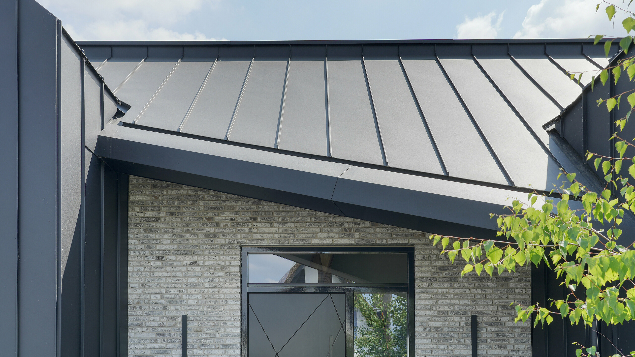 Eingangsbereich eines stilvollen Familienhauses in Veenendaal, Niederlande. Der Eingang wird durch eine schwarze Aluminiumverkleidung hervorgehoben. Das PREFALZ Dach- und Fassadensystem kontrastiert mit dem hellgrauen Naturstein der Fassade. Die große schwarze Tür trägt die Hausnummer 40 und bietet einen eleganten Willkommenspunkt neben der attraktiv gestalteten Gartenlandschaft.