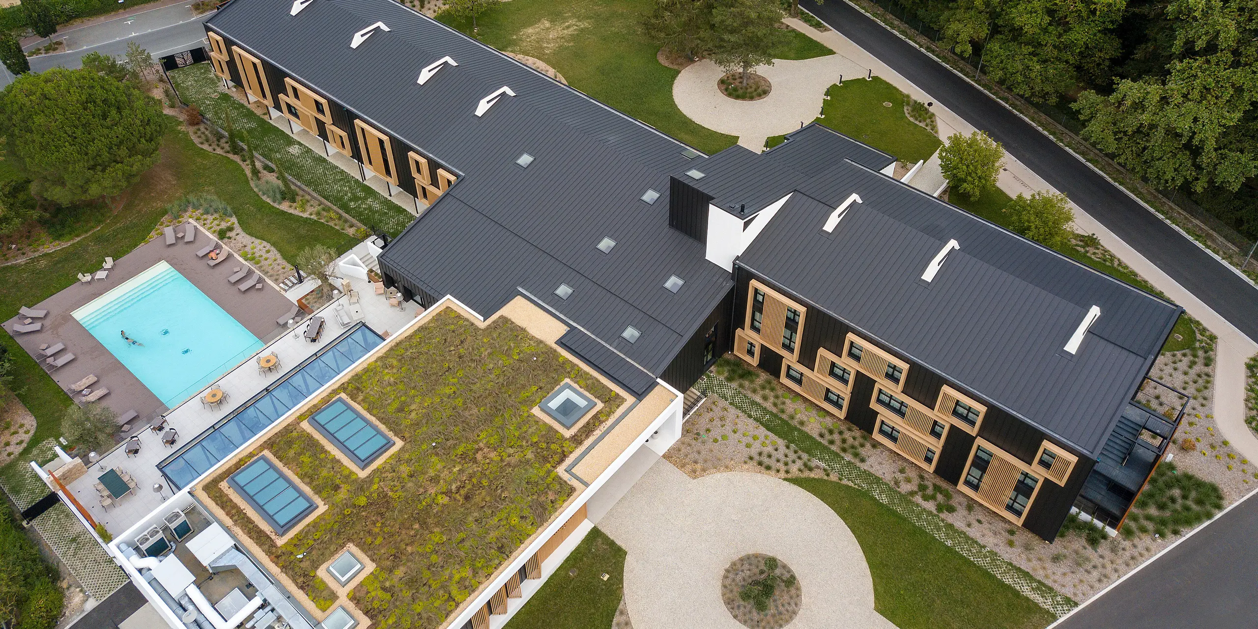 Luftaufnahme auf den Eingangsbereich des Hôtel Parc du Landreau in Les Herbiers, Frankreich. Das Gebäude wurde mit dem PREFALZ Dach- und Fassadensystem in Schwarzgrau verkleidet. Die markanten Holzrahmen um die Fenster ergänzen die moderne Aluminiumverkleidung. Der Außenbereich mit Swimmingpool und teilweise begrüntem Dach spiegelt die nachhaltige Bauweise des Hotels wider.