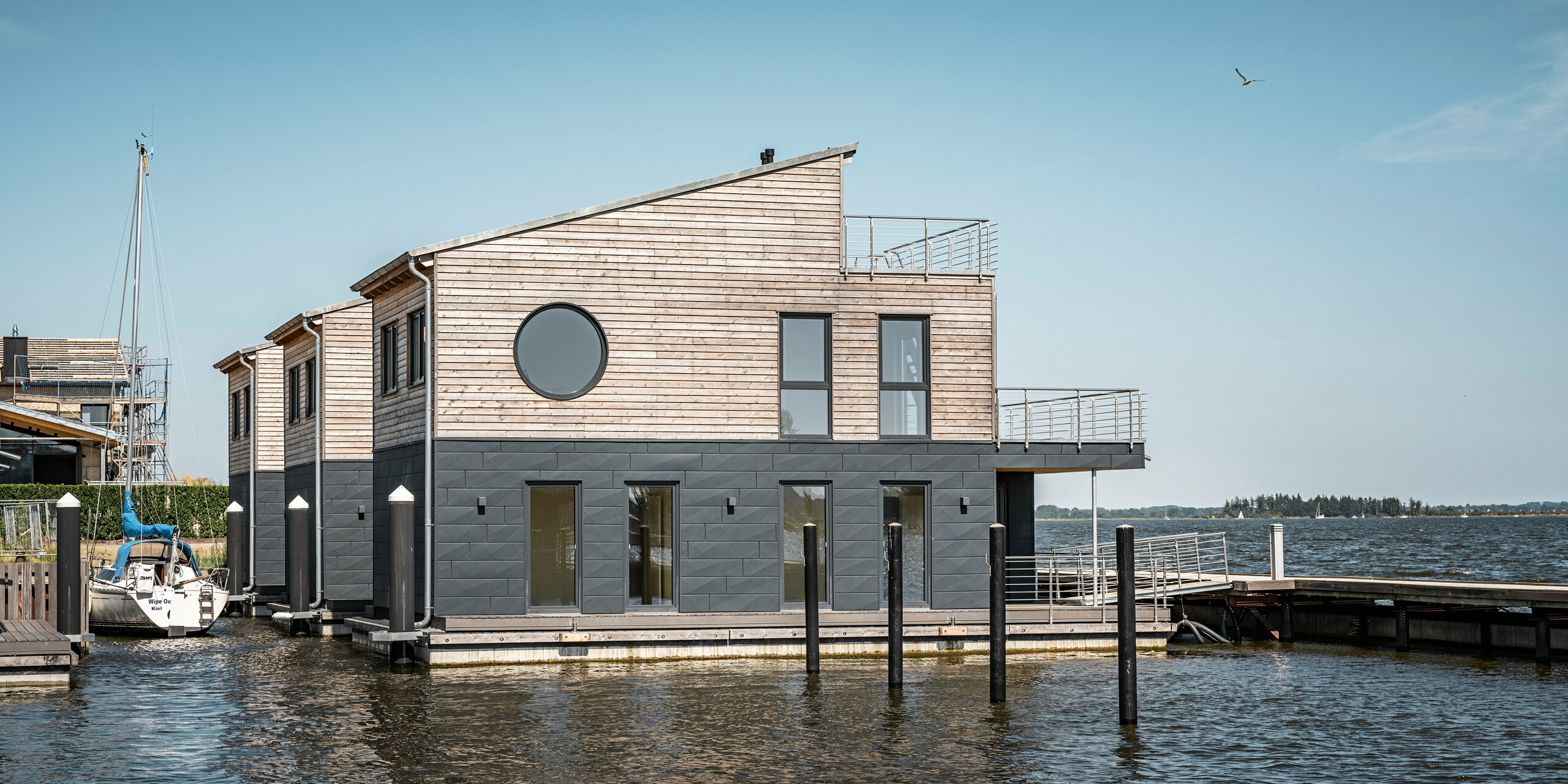 Dieses Bild stellt ein einzelnes, elegant gestaltetes Wasserhaus im Pionierhafen Schleswig in den Vordergrund. Das Wasserhaus steht mit zwei weiteren Häusern in einer Reihe. Hinter dem Haus am linken Bildrand ist ein angelegtes Segelboot zu sehen. Die Fassade der Wasserhäuser kombiniert umweltfreundliches Holz mit langlebigen PREFA Siding.X Aluminiumpaneelen in P.10 Anthrazit, was eine beständige und ästhetische Lösung darstellt. Die ruhige Wasseroberfläche und der offene Blick auf die Schlei runden das Bild eines idyllischen, maritimen Lebensstils ab.