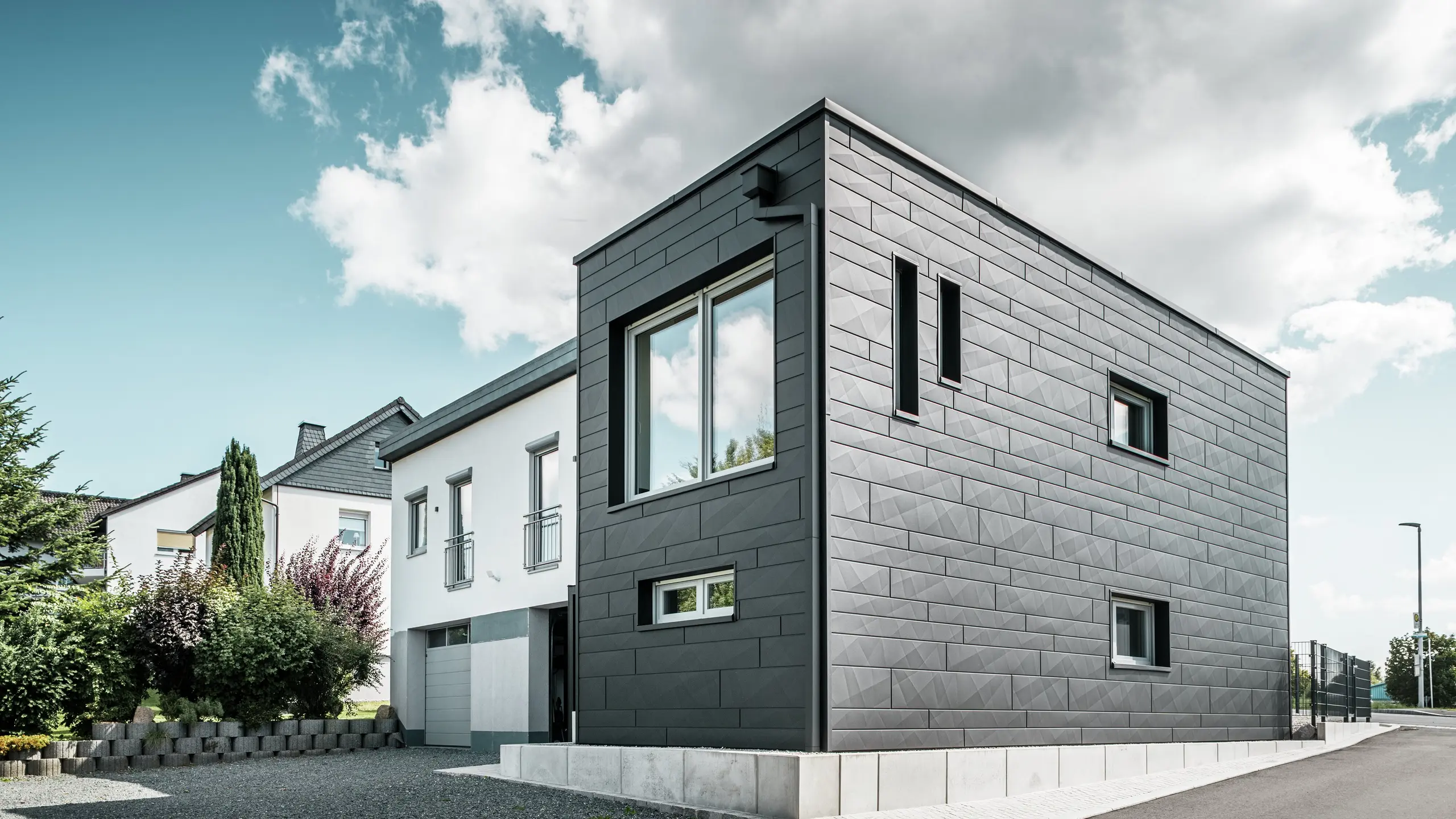 A felújított családi ház stílusos és modern homlokzata Teuschnitzben a világosan strukturált PREFA Siding.X panelekből áll, P.10 fekete színben. A kiváló minőségű fémpanelek geometrikus elrendezése az épületnek markáns, időtálló esztétikát kölcsönöz. Az ablakokat fekete alumínium kompozit panelek keretezik, ami harmonikusan kiegészíti a dizájnt. Az integrált PREFA tetővízelvezető rendszer négyzetes csővel funkcionális és esztétikus megoldást kínál.