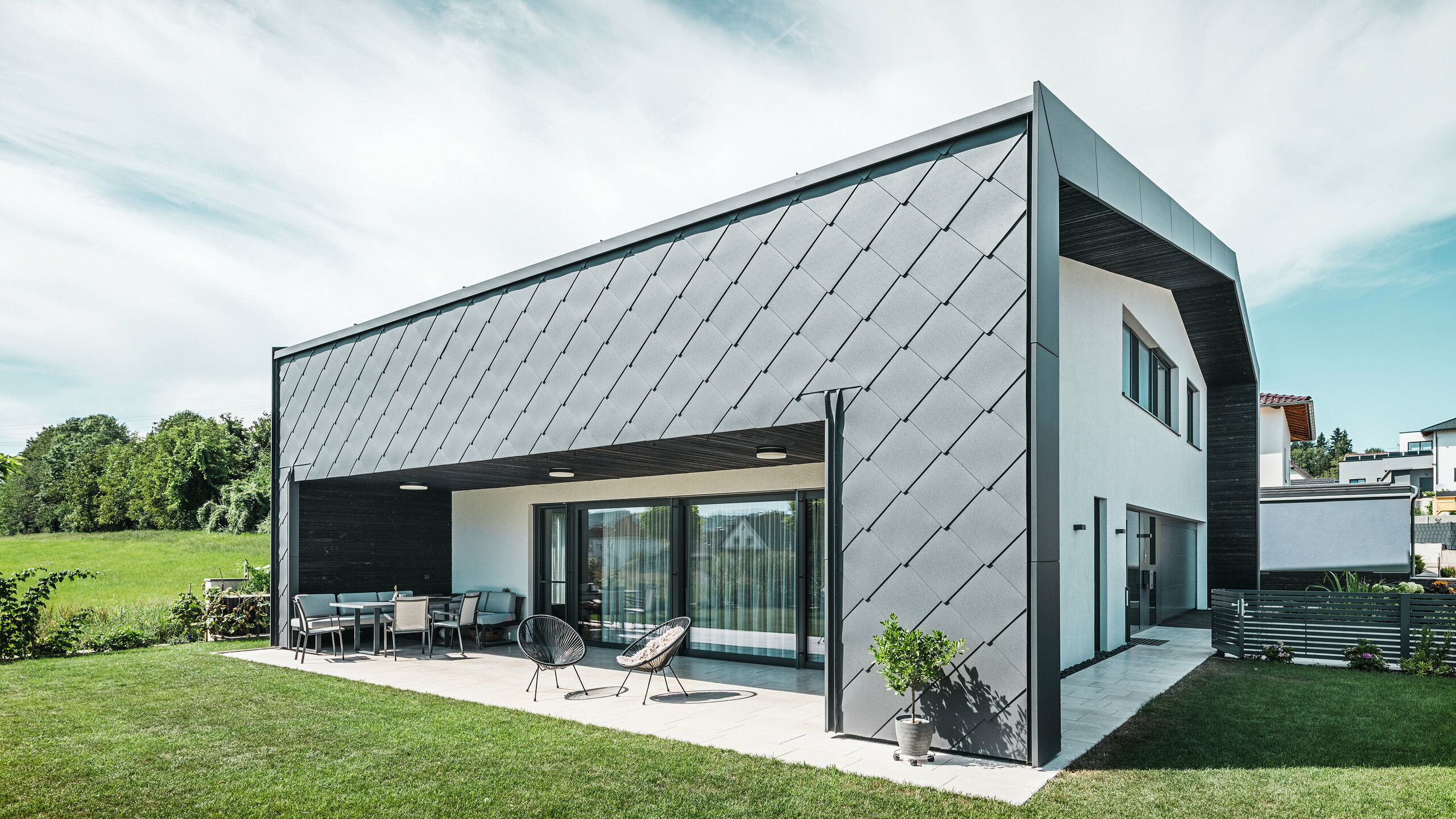 Gepflegter Gartenbereich eines Einfamilienhauses in Aschach. Das Gebäude ist stilvoll eingedeckt mit dunkelgrauen PREFA Aluminium-Wandrauten 44 × 44. Die überdachte Terrasse bietet einen gemütlichen Sitzplatz mit Blick auf die saftig grüne Rasenfläche. Die elegante Aluminiumverkleidung in Kombination mit weißen Putzflächen sorgt für eine moderne und witterungsbeständige Architektur. Große Fensterflächen lassen viel Tageslicht in den Innenraum und schaffen eine offene, lichtdurchflutete Atmosphäre.