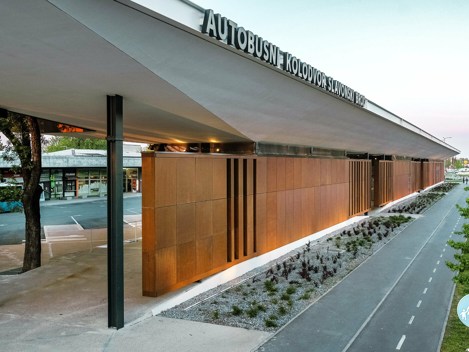 Das Bild zeigt die moderne Bushaltestelle "Autobusni Kolodvor Slavonski Brod" in Kroatien. Die Haltestelle verfügt über eine markante Fassade aus braunen Paneelen, die von schwarzen Säulen gestützt wird. Das weiße Prefalz Dach von PREFA erstreckt sich über die gesamte Struktur und bietet Schutz für die darunter liegenden Bereiche. Im Vordergrund ist ein Fahrradweg zu sehen, der von einer gepflegten Grünfläche und jungen Bäumen flankiert wird. Im Hintergrund sind ein hellblauer Bus und verschiedene Geschäfte zu erkennen.