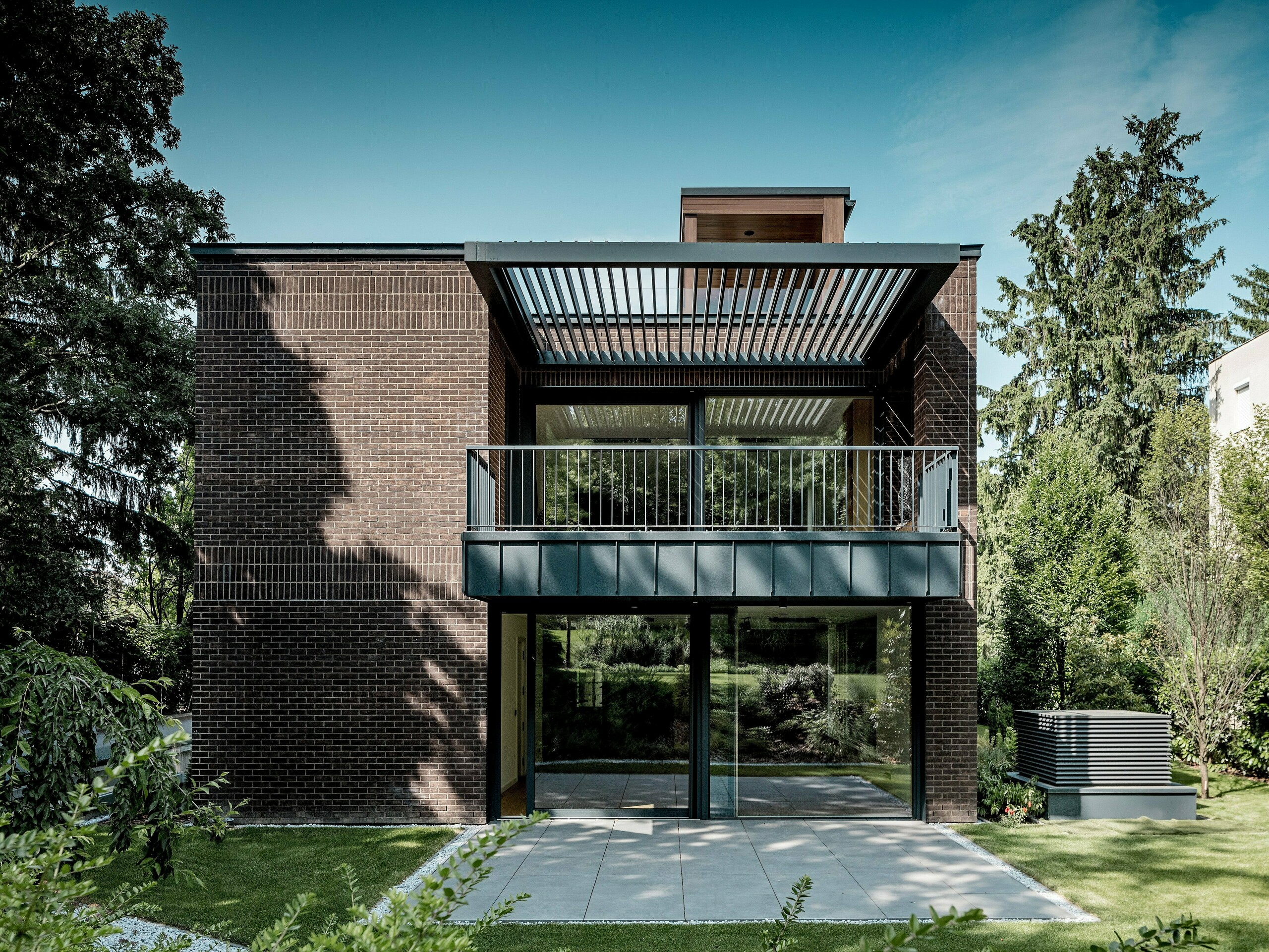 Die Aufnahme zeigt die Gartenseite der modernen Mehrgenerationen‑Villa in Budapest mit ihrem klar strukturierten Fassadenaufbau aus dunkelrotem Klinker und PREFALZ Aluminium in P.10 Anthrazit. Der große Balkon mit filigraner Metallkonstruktion und darüber liegenden Lamellen erzeugt ein spannendes Spiel aus Licht und Schatten. Bodentiefe Glasfronten öffnen den Innenraum zur grünen Gartenlandschaft. Die präzise ausgeführten Aluminiumdetails unterstreichen die Langlebigkeit, Witterungsbeständigkeit und architektonische Klarheit der PREFA Lösungen, die dem Gebäude einen markanten, zeitlosen Ausdruck verleihen.