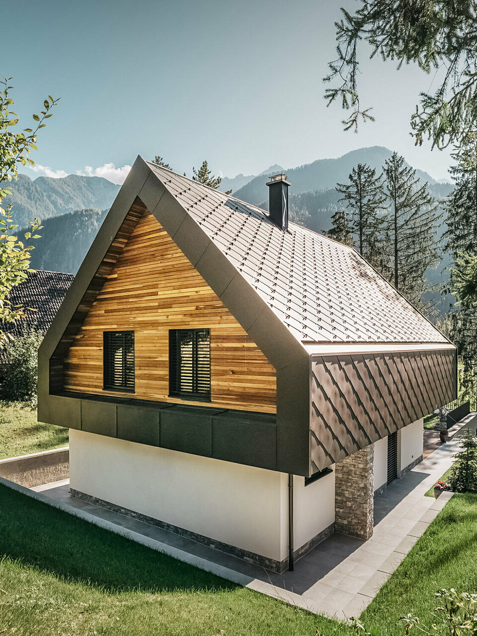 Ein modernes Chalet in Slowenien mit einer Kombination aus traditionellen und zeitgenössischen Architekturelementen, eingebettet in eine alpine Landschaft. Das Haus weist eine hölzerne Fassade mit geschlossenen Fensterläden auf, ein charakteristisches steiles Satteldach mit nussbraunen Dach- und Wandrauten und einen Schornstein. Das Anwesen ist umgeben von reichlich Grünflächen, Bäumen und bietet einen klaren Blick auf die entfernten Berge und ein offenes Tal