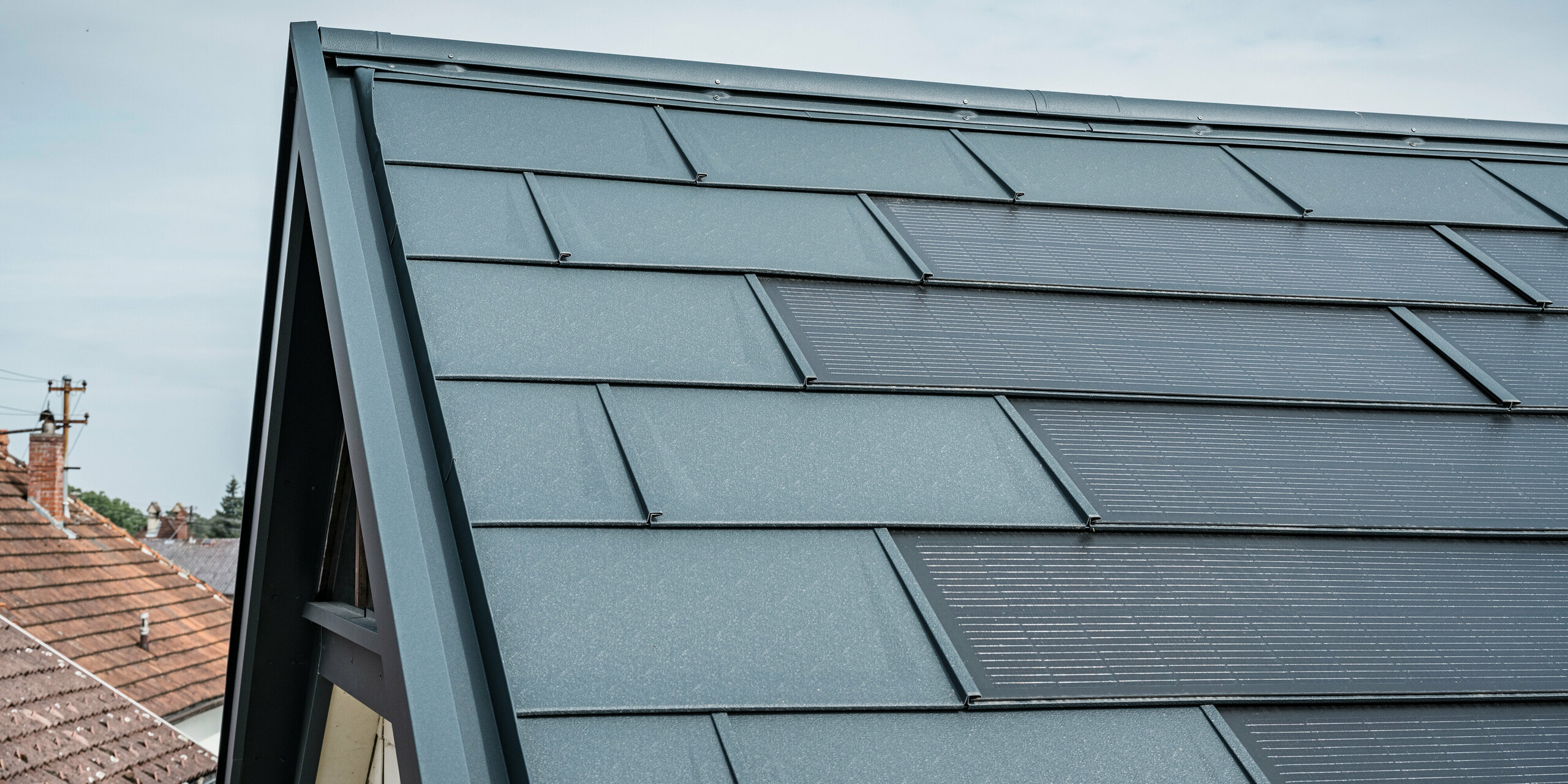 Az innovatív PREFA napelemes tetőpanellel felszerelt ház tetejének részletes nézete. A beépített fotovoltaikus cellákkal ellátott tetőpanelek elegáns antracit színűek. A homogén felület zökkenőmentesen illeszkedik a tetőbe, modern és letisztult megjelenést kölcsönözve. Az innovatív tetőrendszer hatékony energiafelhasználást biztosít anélkül, hogy az esztétika rovására menne.