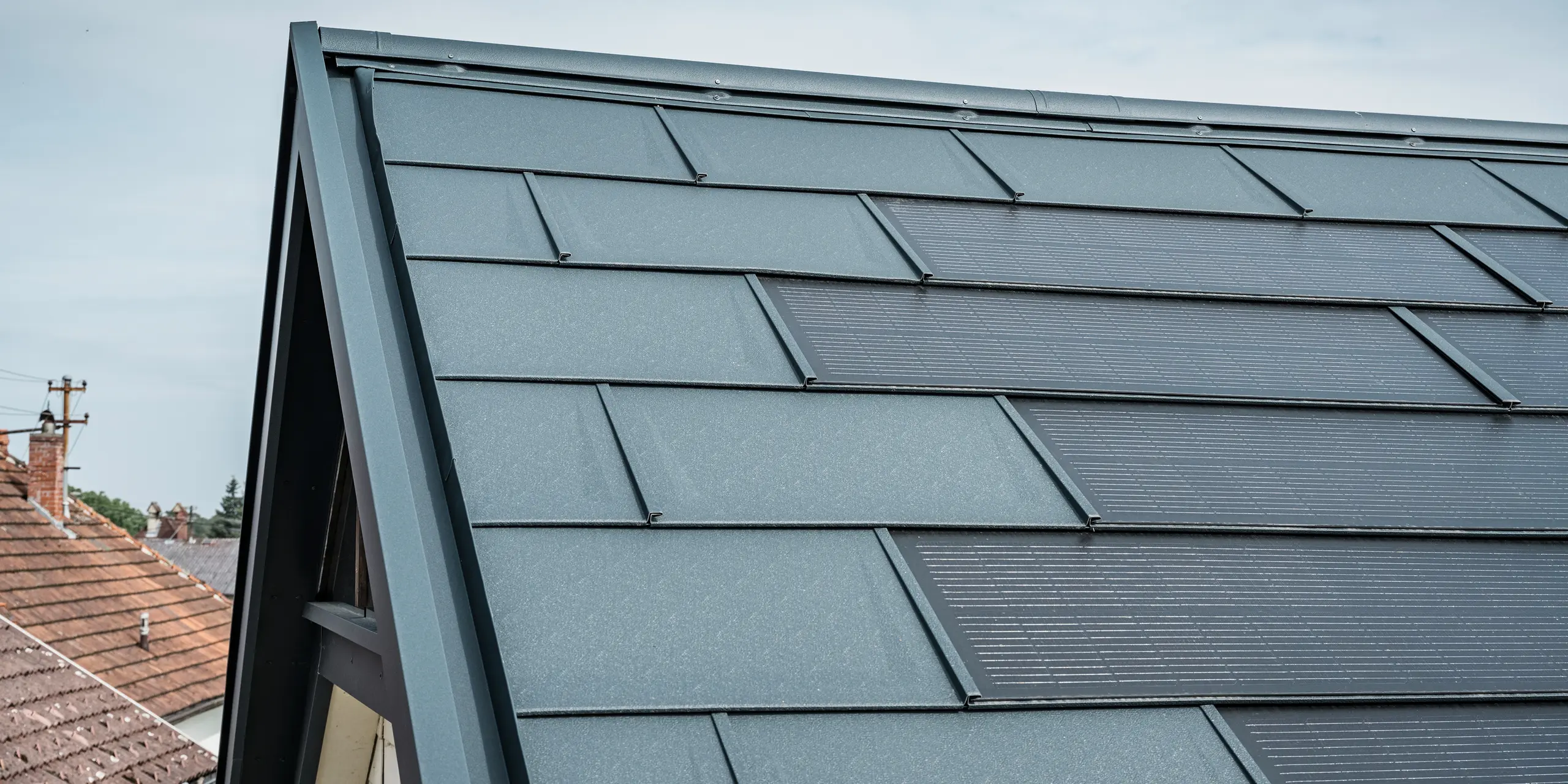 Az innovatív PREFA napelemes tetőpanellel felszerelt ház tetejének részletes nézete. A beépített fotovoltaikus cellákkal ellátott tetőpanelek elegáns antracit színűek. A homogén felület zökkenőmentesen illeszkedik a tetőbe, modern és letisztult megjelenést kölcsönözve. Az innovatív tetőrendszer hatékony energiafelhasználást biztosít anélkül, hogy az esztétika rovására menne.