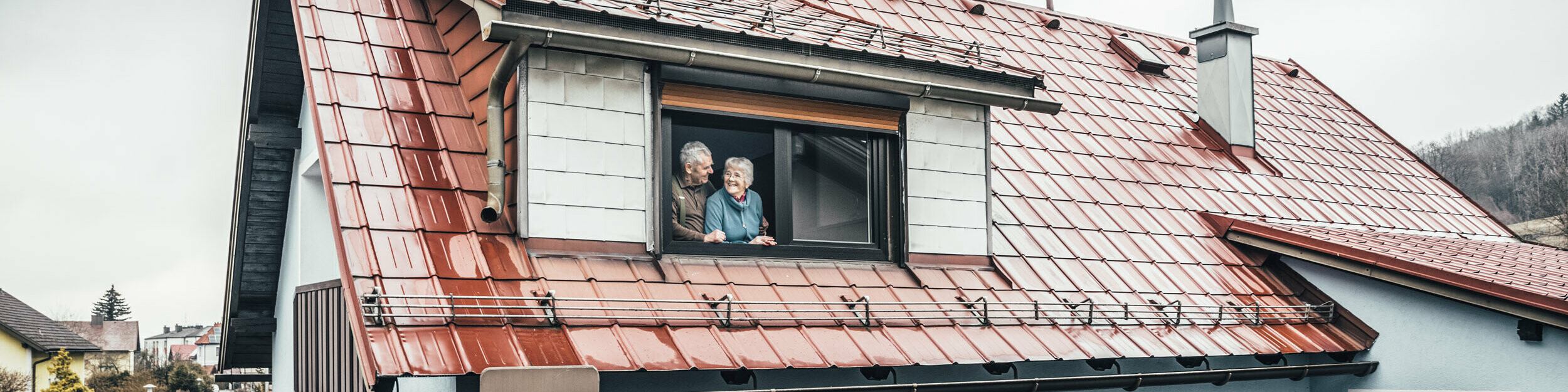 Anna und Heinrich stehen in ihrem Eigenheim - eingedeckt mit der PREFA Dachplatte in der Farbe Ziegelrot - am offenen Fenster und sehen sich glücklich und zufrieden an. 