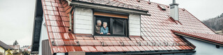 Anna und Heinrich stehen in ihrem Eigenheim - eingedeckt mit der PREFA Dachplatte in der Farbe Ziegelrot - am offenen Fenster und sehen sich glücklich und zufrieden an. 