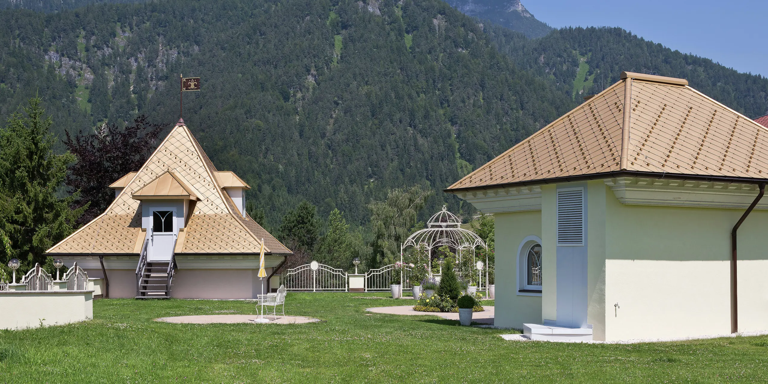 Gesamtansicht zweier kleiner Anbauten an ein Einfamilienhaus in Tirol. Die beiden goldenen Dächer sind mit PREFA Dachrauten in Mayagold eingedeckt. Im Vordergrund ein gepflegter Garten, im Hintergrund die beeindruckende Bergkulisse.