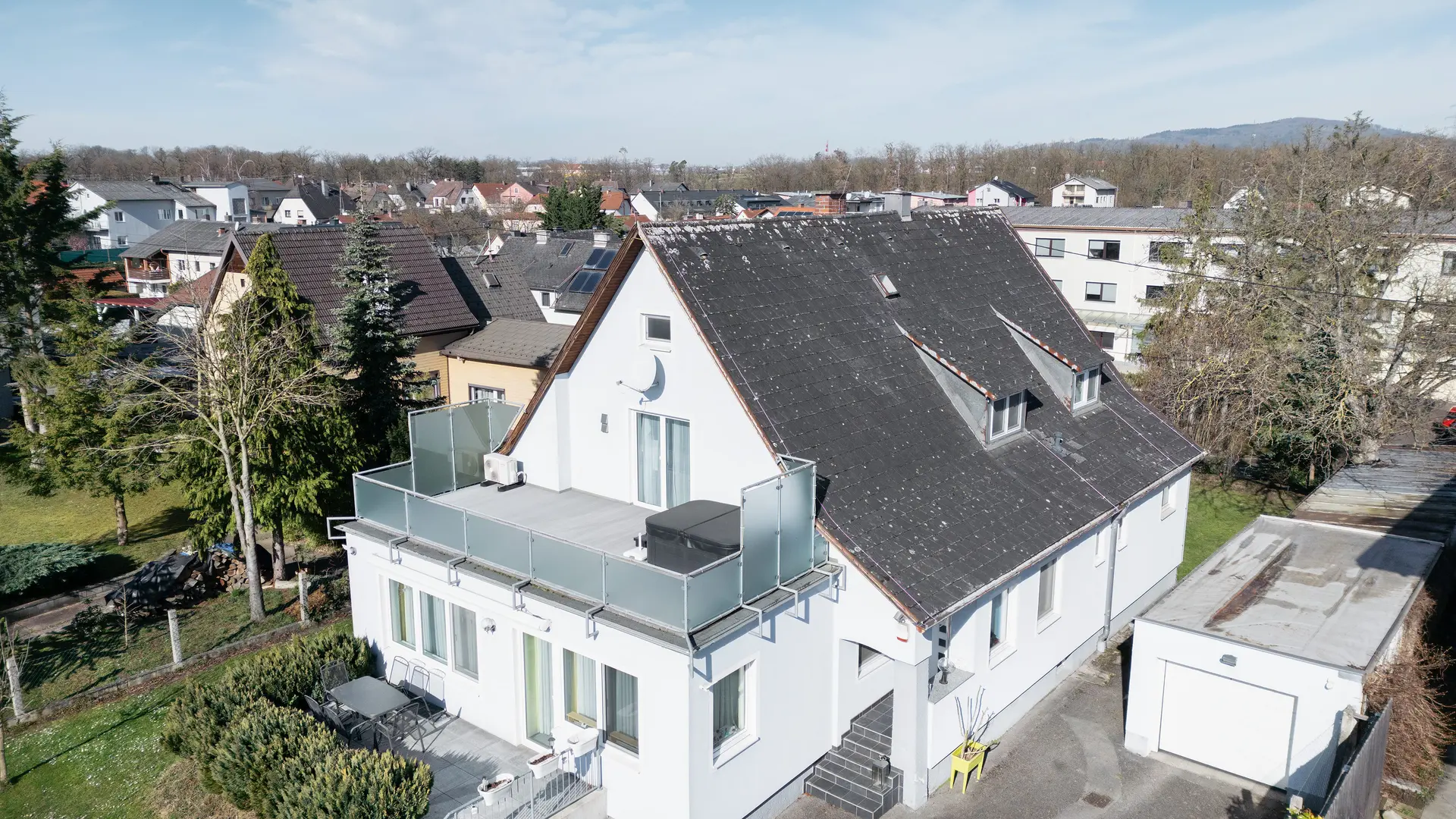 Madártávlatból egy régi, szennyeződéssel és zuzmóval borított eternit tetővel rendelkező épület. A tetőn nem látható napelem-rendszer. Az előtérben látható erkély üvegkorláttal rendelkezik, és az épület felső emeletén található.