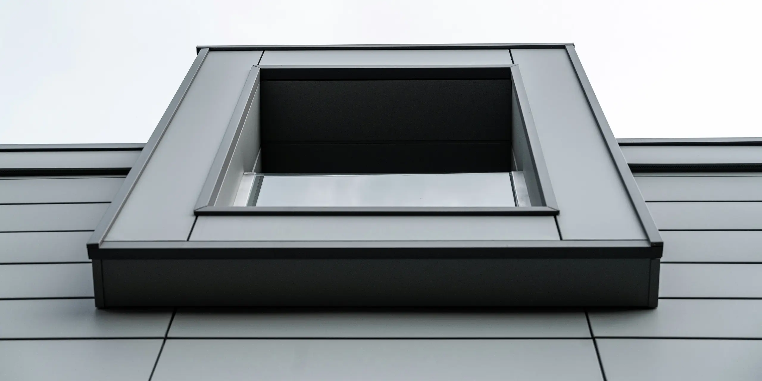 Nahaufnahme von unten auf ein auskragendes Fenster einer Wohnanlage in Faloppio, Italien. Die klare, geometrische Form des Fensters in Kombination mit der langlebigen PREFA Siding Aluminium-Fassade unterstreicht die moderne Architektur und das minimalistische Design des Gebäudes. 