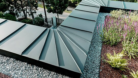 Die Aufnahme zeigt eine detaillierte Nahansicht der Attika‑Ausführung der Budapester Mehrgenerationen‑Villa, verkleidet mit PREFALZ Aluminium in P.10 Anthrazit. Die fächerförmig angeordneten Scharen laufen präzise an der Gebäudekante zusammen und demonstrieren die hohe Formbarkeit und Verarbeitungsqualität der PREFA Aluminiumprodukte. Das anthrazitfarbene Metall bildet einen eleganten Kontrast zum begrünten Dachbereich mit Kiesbett und blühenden Pflanzen. Die Szene betont die Langlebigkeit, Witterungsbeständigkeit und technische Raffinesse der PREFA Lösung.