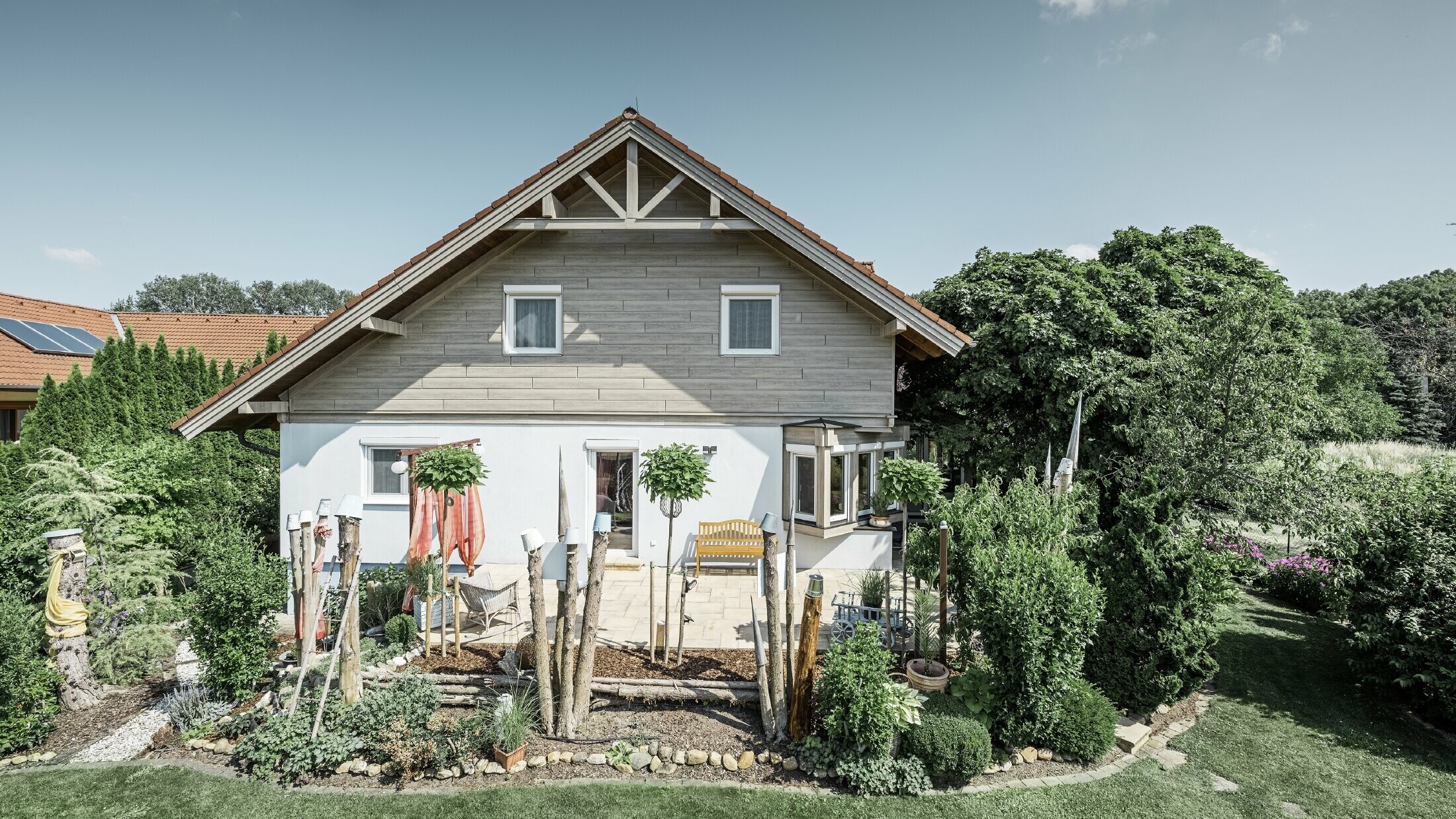 Homlokzatát felújított családi ház, modern, szürke-bézs színű, fa hatású homlokzati burkolattal, vakolt homlokzattal, nyeregtetővel és gondozott kerttel.