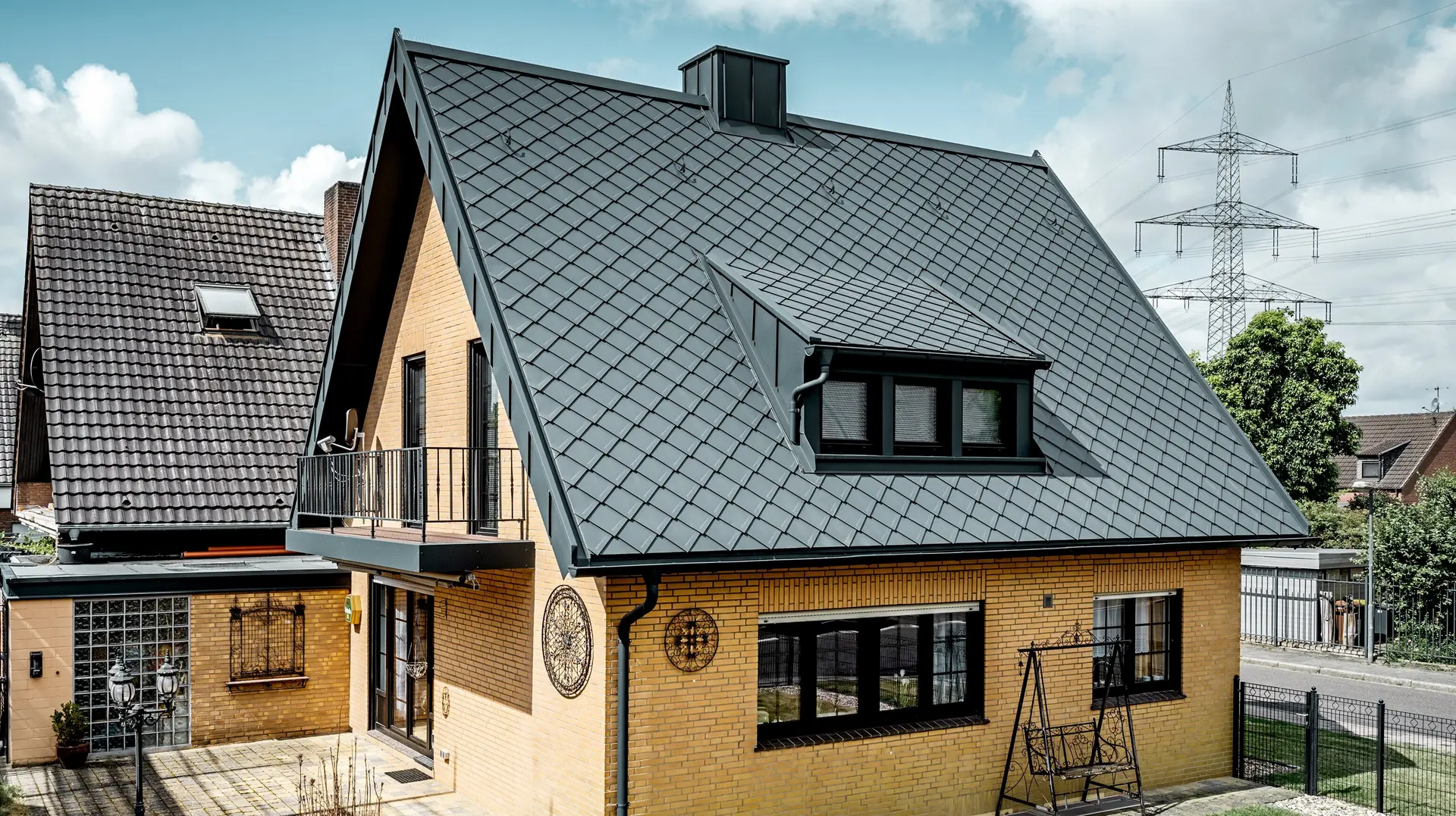 Sárga téglából épült családi ház a tetőfelújítás után, PREFA tetőfedő rombusz 29 × 29 grafitszürke színben; új tetőfedés, meredek nyeregtető rombuszmintával, beépített tetőablakkal és kéménnyel, terasszal és kerttel az előtérben.