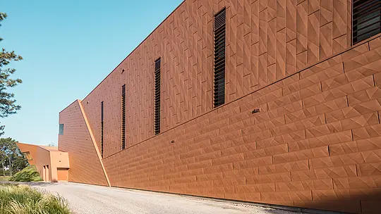 Dynamisch gefaltete Fassade des Trinkwasserpumpwerks WML in Lottum mit präzise gesetzten, schmalen Fensteröffnungen als architektonische Eyecatcher. Die markante PREFA Siding.X Aluminiumfassade in warmer Rostoptik erzeugt ein lebendiges Spiel aus Licht, Schatten und Geometrie entlang der klaren Baukörperkante. Die außergewöhnliche Struktur verbindet technische Funktion mit skulpturaler Ästhetik. Die leichten, langlebigen und vollständig recycelbaren PREFA Aluminium-Paneele überzeugen durch hohe Witterungsbeständigkeit, Formstabilität und eine nachhaltige, wartungsarme Gebäudehülle.