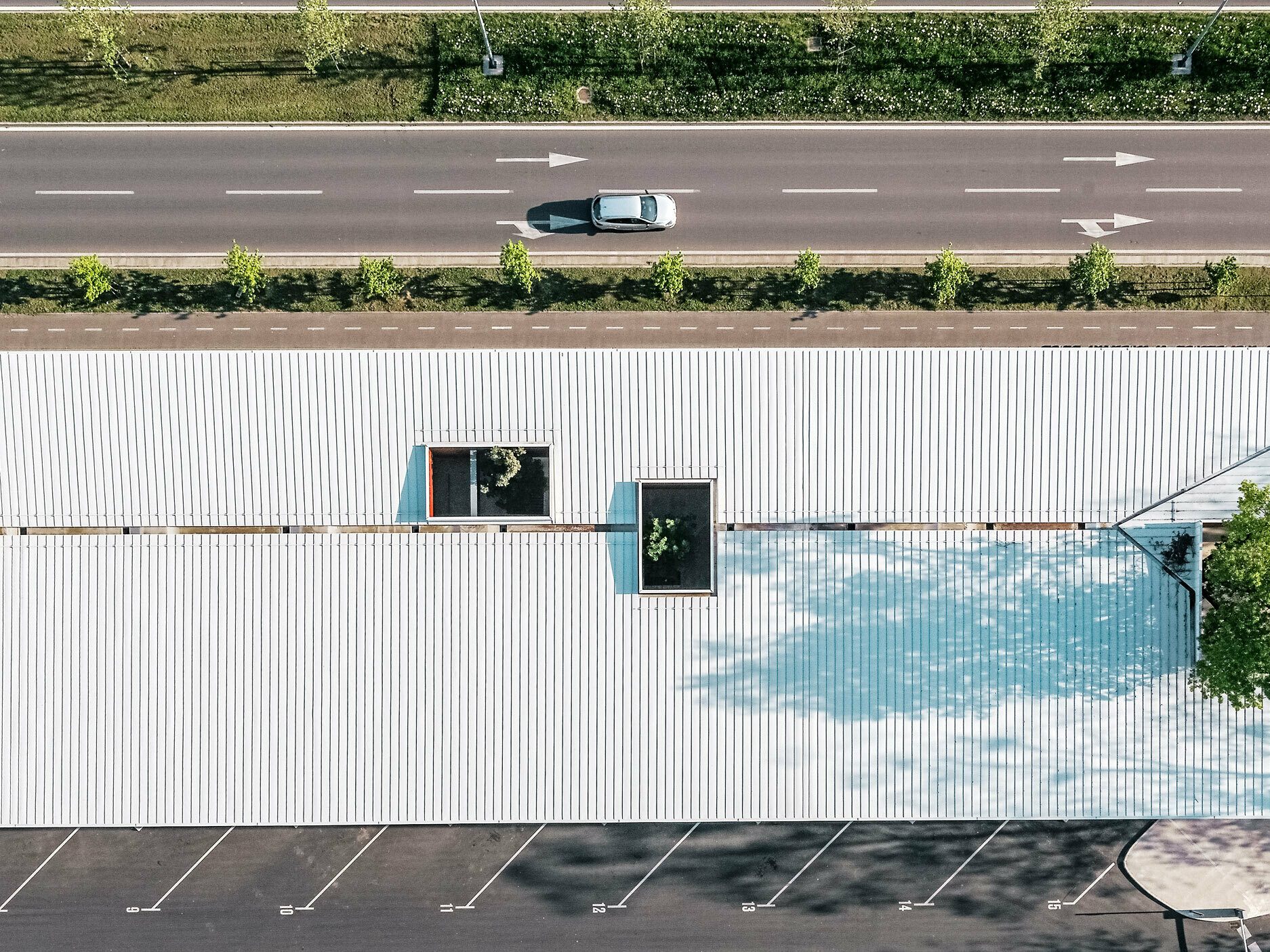 Das Bild zeigt eine Luftaufnahme der Bushaltestelle in Kroatien mit einem weißen Prefalz Dach von PREFA. Das lange Gebäude hat mehrere rechteckige Dachfenster, die in verschiedenen Positionen angeordnet sind und das grüne Laub von Bäumen durchscheinen lassen. Ein kleinerer Parkplatz ist im Vordergrund zu sehen, während sich im Hintergrund eine zweispurige Straße mit einer Linie von Bäumen entlangzieht.