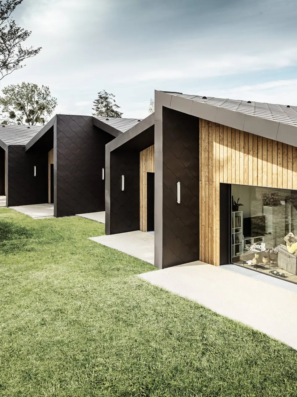 Das Bild zeigt eine Reihe moderner, minimalistisch gestalteter Gebäude mit einer Fassade aus PREFA 44 × 44 Dach- und Wandrauten in Walnussbraun. Die Rautenstruktur der Verkleidung wird harmonisch durch warme Holzelemente ergänzt, die die Eingangsbereiche und großen Glasflächen einrahmen. Die Gebäude befinden sich in einer grünen, sorgfältig gestalteten Landschaft, die eine ruhige und angenehme Atmosphäre schafft. Dieser Entwurf vereint Funktionalität mit ästhetisch ansprechender Architektur.