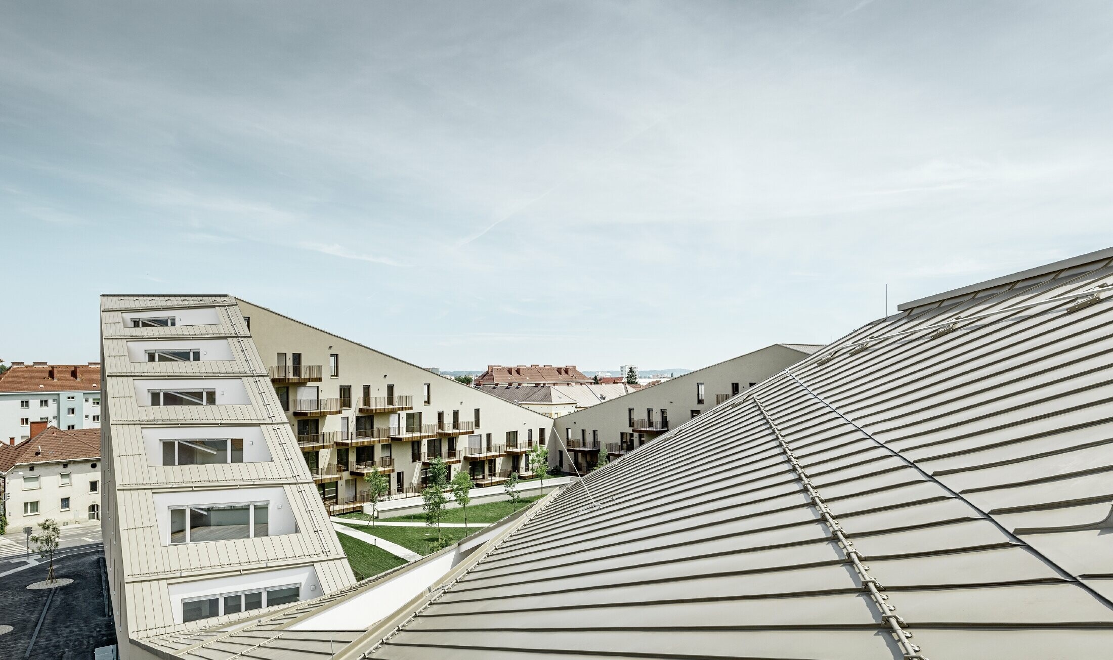 hosszasan elnyúló lakótelep Grazban, alacsony hajlású tetősíkokkal, beépített teraszokkal és nagy üvegfelületekkel, egyedi, bronz metál Prefalz korcolt lemezfedéssel