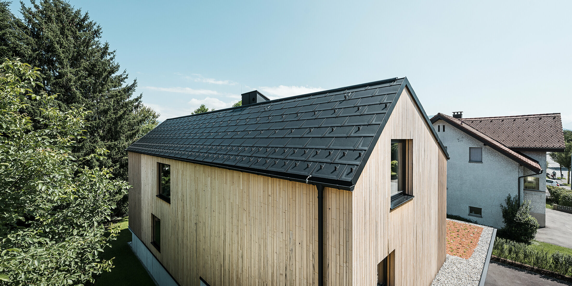 Ein modernes Einfamilienhaus in Lustenau besticht durch die Kombination einer freundlichen Holzfassade mit robusten PREFA Aluminiumprodukten. Das Satteldach wird von der PREFA Dachplatte R.16 mit Schneestoppern sowie der passenden Dachentwässerung in Form von Hängerinne und Ablaufrohr geschmückt. Alle Produkte sind einheitlich im Farbton Schwarz P.10 gehalten und harmonieren perfekt mit der vertikalen Holzfassade.
