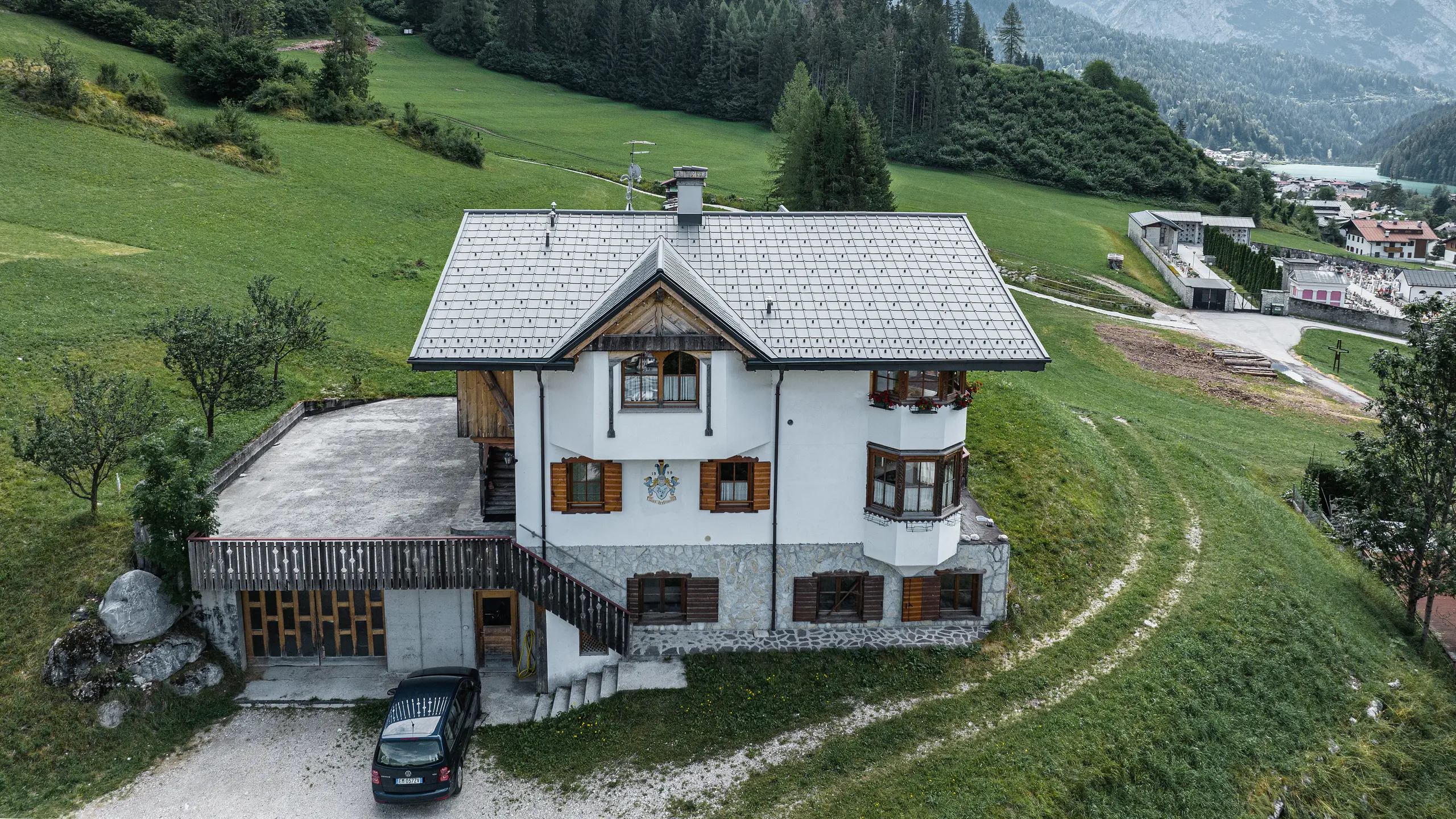 Madártávlatból az Auronzo di Cadore-i hagyományos hegyi kunyhó bejárata a PREFA P.10 világosszürke színű alumíniumcserepekkel történő tetőfelújítás után. A jellegzetes alpesi bájjal és modern bádogtetővel rendelkező épület harmonikusan illeszkedik a buja zöld tájba és a háttérben lévő lenyűgöző hegyi panorámába, a funkcionalitás és a regionális építészet sikeres kombinációját mutatva.