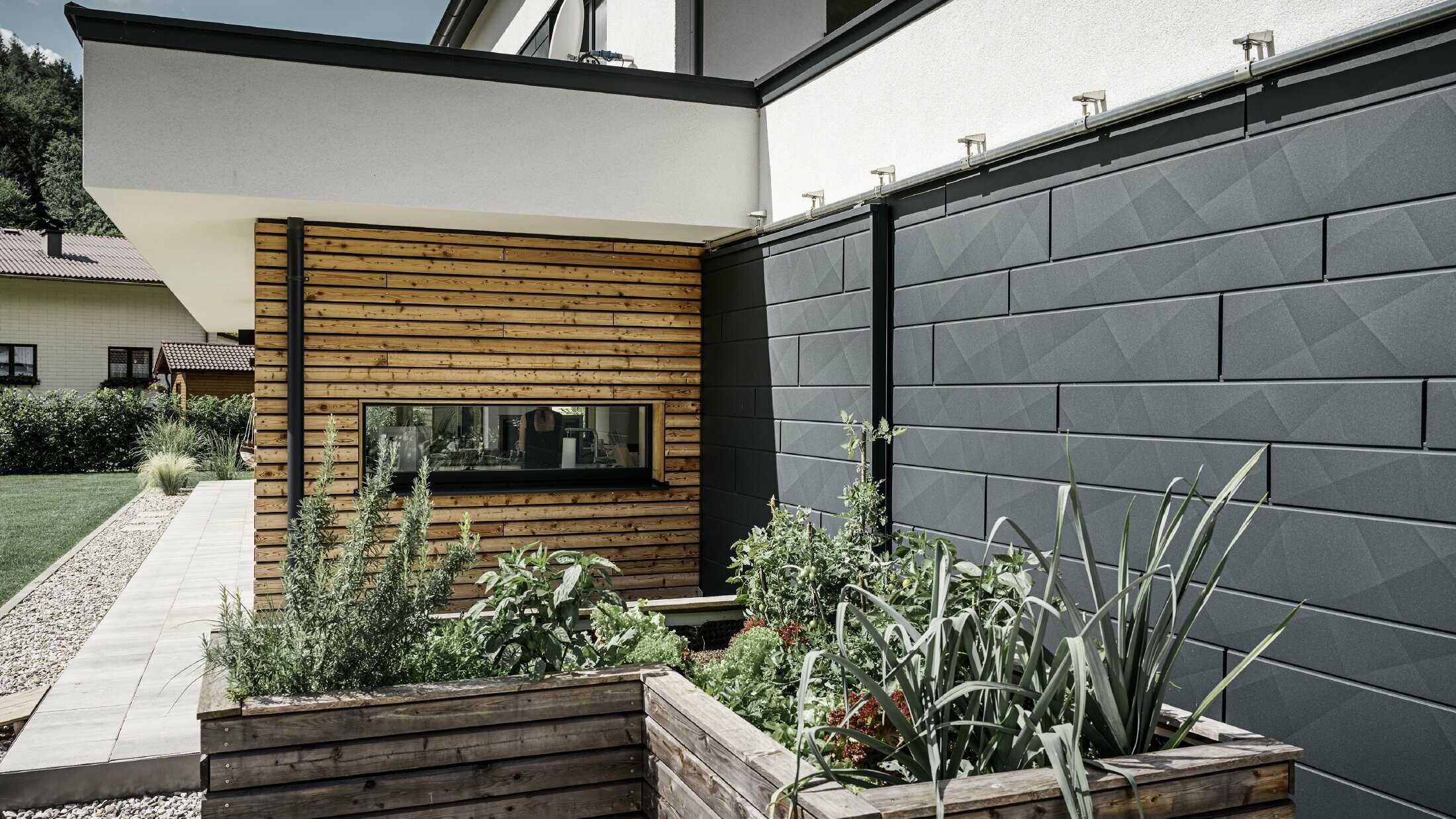 A képen egy családi ház közelképe látható, amelyen a PREFA Siding.X P.10 antracit színű homlokzati burkolat áll a középpontban. A sötét, jellegzetes ráncos felületű homlokzatburkoló panelek vonzó vizuális kontrasztot alkotnak az élénk, fával burkolt részekkel és a környező zöldterületekkel. Az alumínium homlokzat előtt magaságyások találhatók, amelyekbe különböző növényeket és cserjéket ültettek, ami hangsúlyozza a ház természetes és fenntartható megjelenését. A modern anyagok kombinációja és a kertbe való integráció egy olyan kortárs építészeti képet közvetít, amely harmonikusan illeszkedik a környezetbe.
