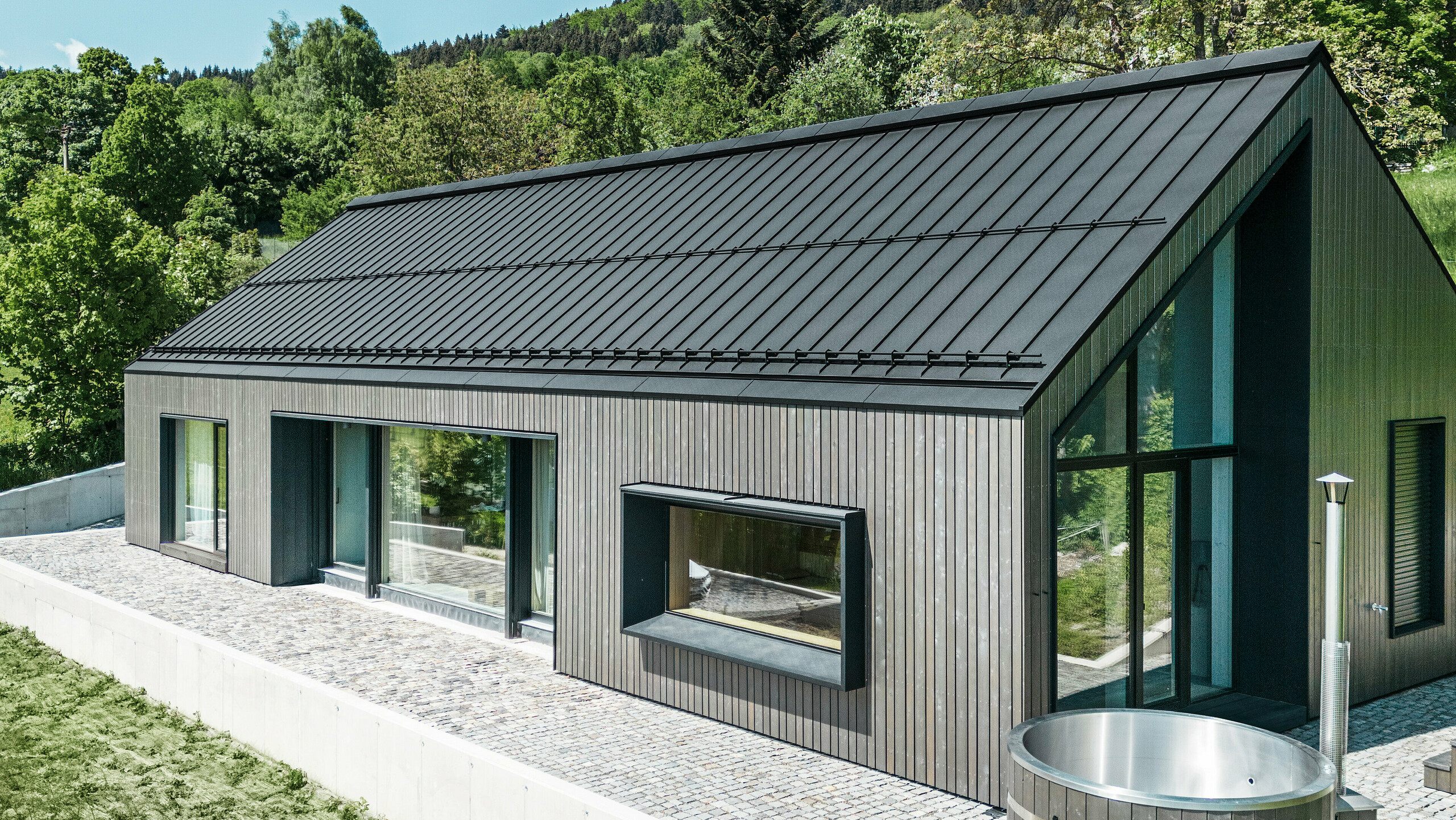 Ein modernes Holzmassivhaus in Kuncice (CZ) wurde mit großen Fensterflächen ausgestattet. Das dunkle Holzmassivhaus verfügt über ein Dachsystem aus PREFALZ in der Farbe P. 10 Schwarz. Im hinteren Außenbereich steht ein Whirlpool mit einer Holzverkleidung.