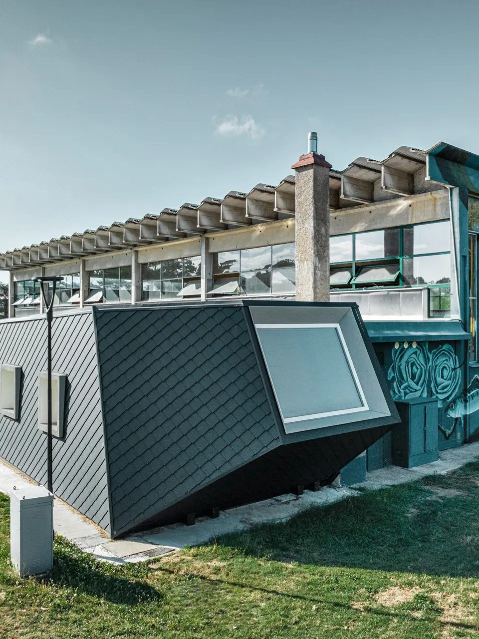 Die Turnhalle Rodriguez und ihr neuer Zubau mit der Rautenschuppenhaut aus PREFA Aluminium von der Seite; der Rasen erstreckt sich davor und zur Seite, im Hintergrund ist ein Teil eines Sportplatzes zu sehen.