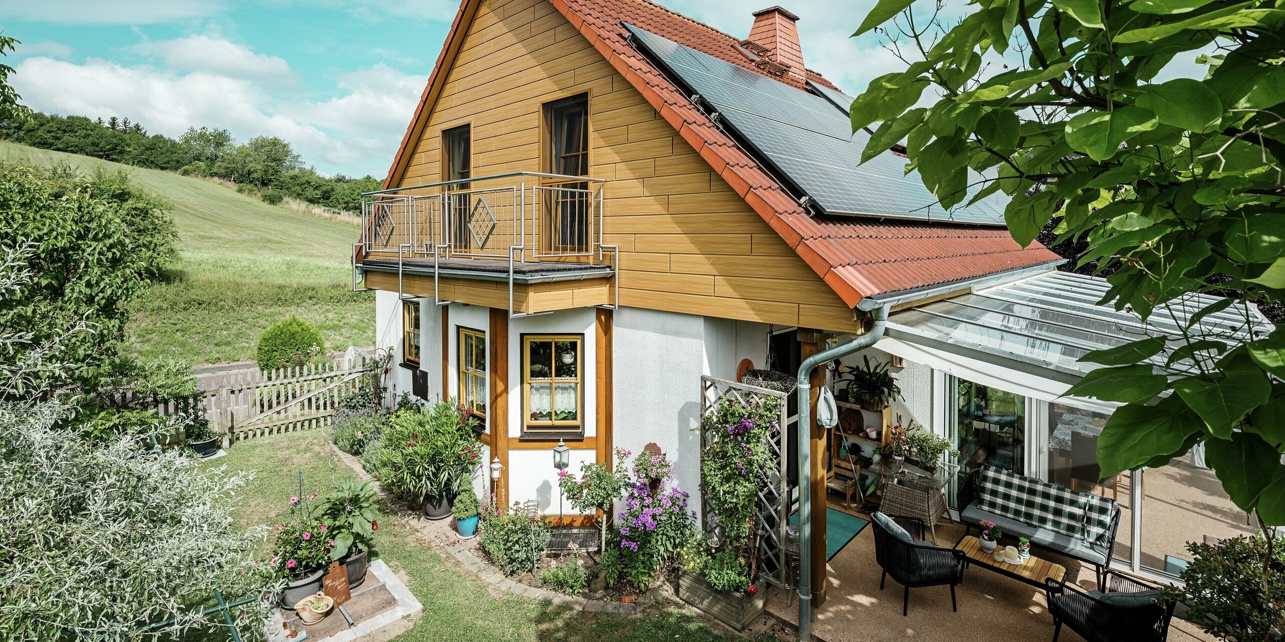 Dieses Bild zeigt ein charmantes Einfamilienhaus in Blieskastel, dessen frisch sanierter Giebel mit PREFA Sidings in Eiche Natur sofort ins Auge fällt. Die warme Holzoptik aus langlebigem Aluminium harmoniert beeindruckend mit dem roten Ziegeldach und dem großzügigen Wintergarten. Umgeben von üppigem Grün und liebevoll angelegten Blumenbeeten entsteht eine idyllische, lebendige Szene. Die robuste PREFA Fassade verleiht dem Haus nicht nur einen natürlichen Look, sondern überzeugt auch durch Witterungsbeständigkeit, Farbtreue und nahezu wartungsfreie Langlebigkeit.