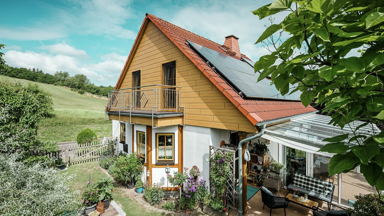 Ez a kép egy bájos családi házat mutat Blieskastelben, amelynek frissen felújított oromzata a világos tölgy PREFA Siding burkolattal azonnal magára vonja a tekintetet. A tartós alumíniumból készült meleg fahatás lenyűgözően harmonizál a vörös cseréptetővel és a tágas télikerttel. A dús zöld növényzet és a gondosan kialakított virágágyások idilli, életteli jelenetet teremtenek. A PREFA-val végzett időtálló homlokzatfelújítás nemcsak természetes megjelenést kölcsönöz a háznak, hanem időjárás-állóságával, színtartósságával és a szinte karbantartásmentes tartósságával is meggyőz.