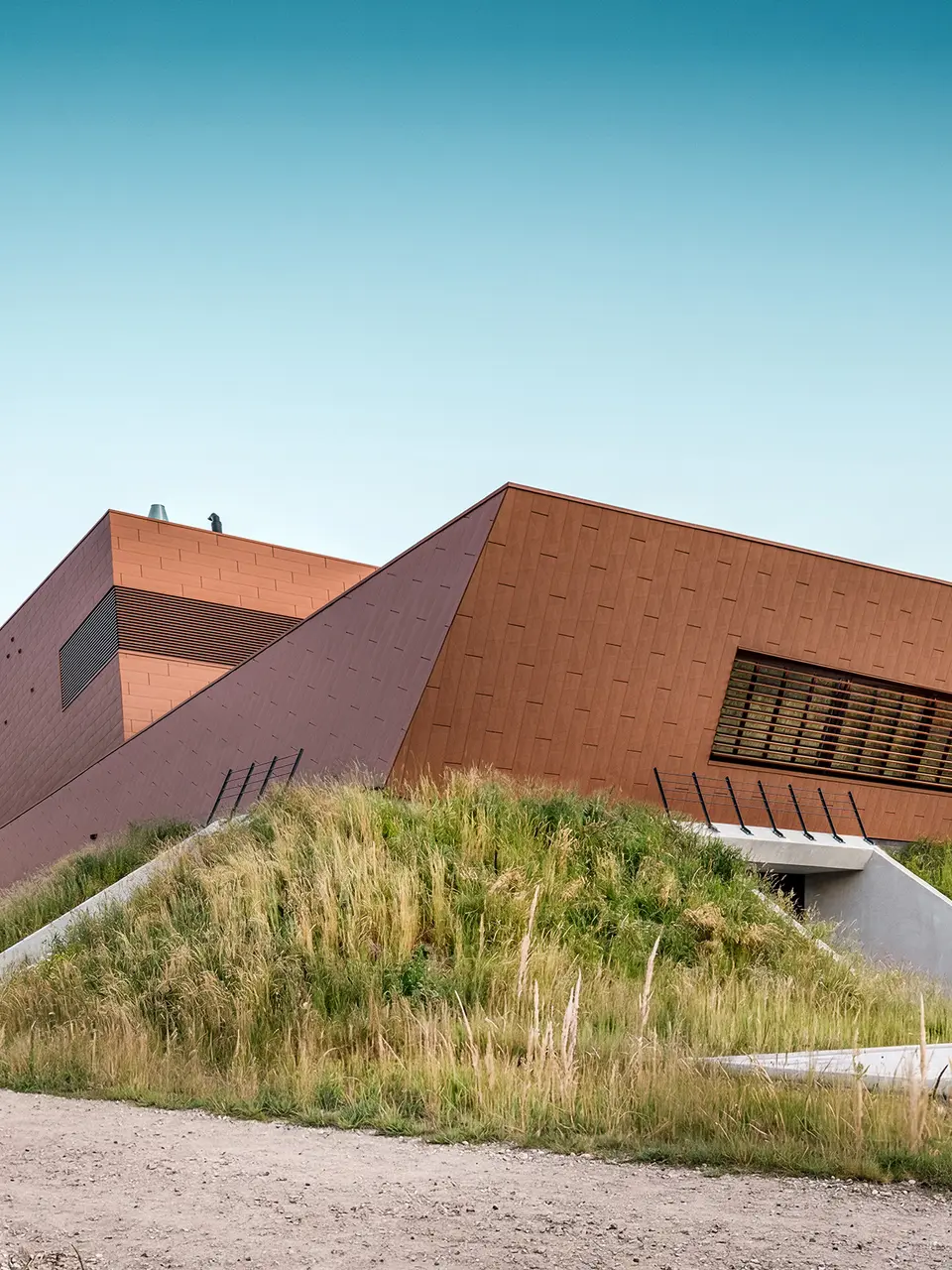 Architektonisch expressives Trinkwasserpumpwerk WML in Lottum mit skulptural gefalteten Baukörpern, eingebettet in eine naturnahe Böschungslandschaft. Die markante PREFA Siding.X Aluminiumfassade in warmer Rostoptik zieht mit ihrer facettierten, lebendigen Oberfläche alle Blicke auf sich und spielt eindrucksvoll mit Licht und Schatten. Horizontale Lamellen und präzise Linienführung betonen die dynamische Geometrie. Die leichten, langlebigen und vollständig recycelbaren PREFA Aluminium-Paneele stehen für höchste Witterungsbeständigkeit, nachhaltige Bauweise und eine wartungsarme Gebäudehülle.